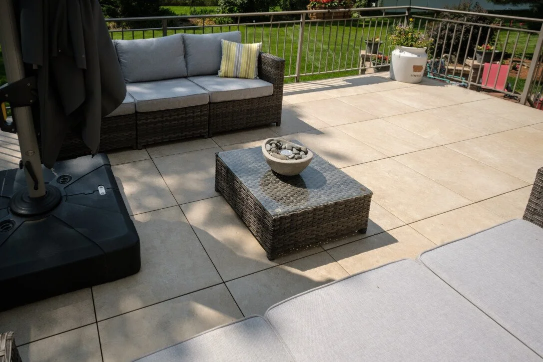 Outdoor patio with wicker furniture including a sofa with light gray cushions, a glass-top coffee table with decorative stones, and a corner section of a cushioned lounge chair. There is a potted plant and garden in the background, surrounded by a metal railing.