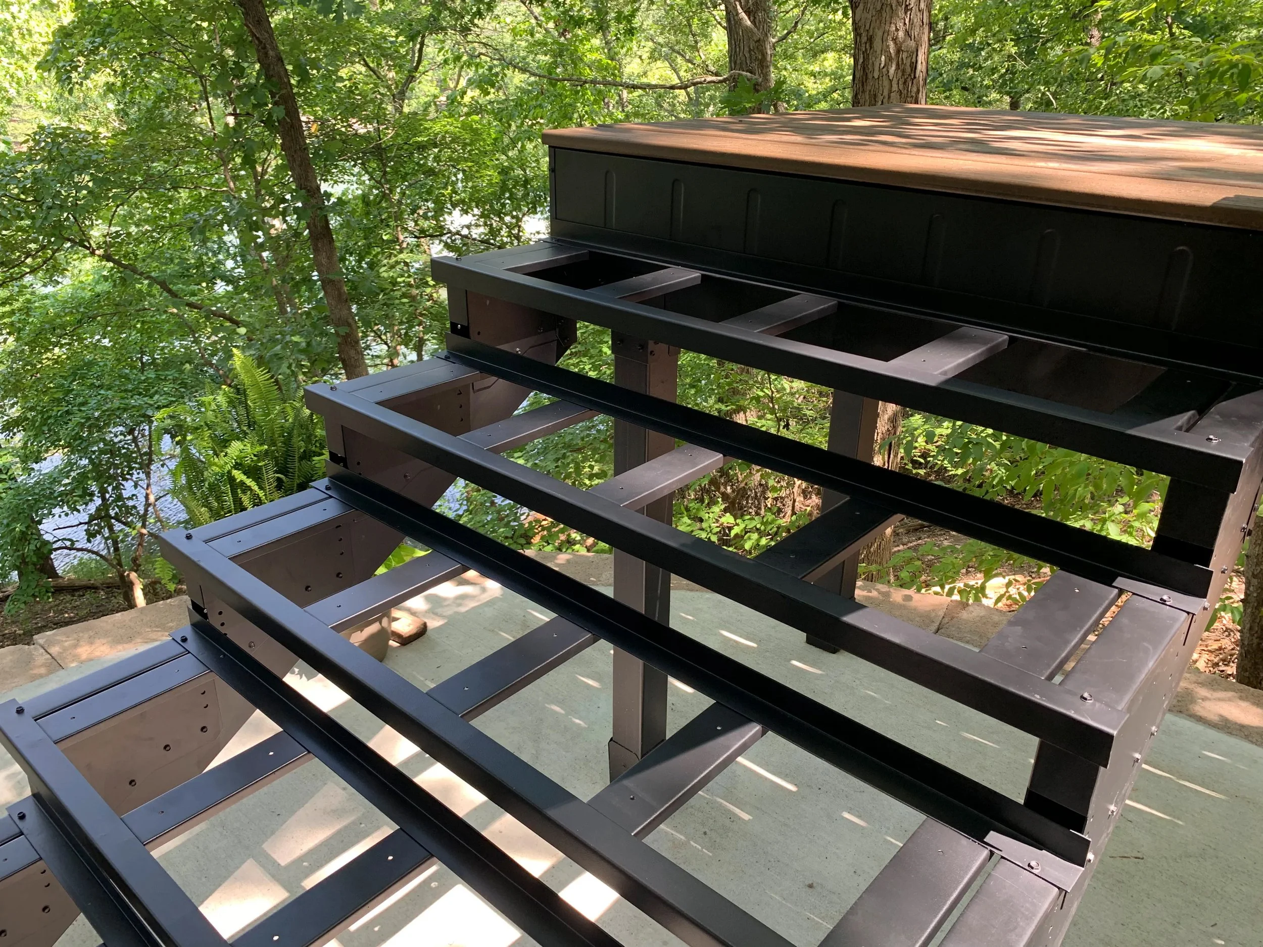 Partially assembled black metal outdoor staircase or deck railing with steps, set on concrete, surrounded by lush green trees and foliage, under bright sunlight.