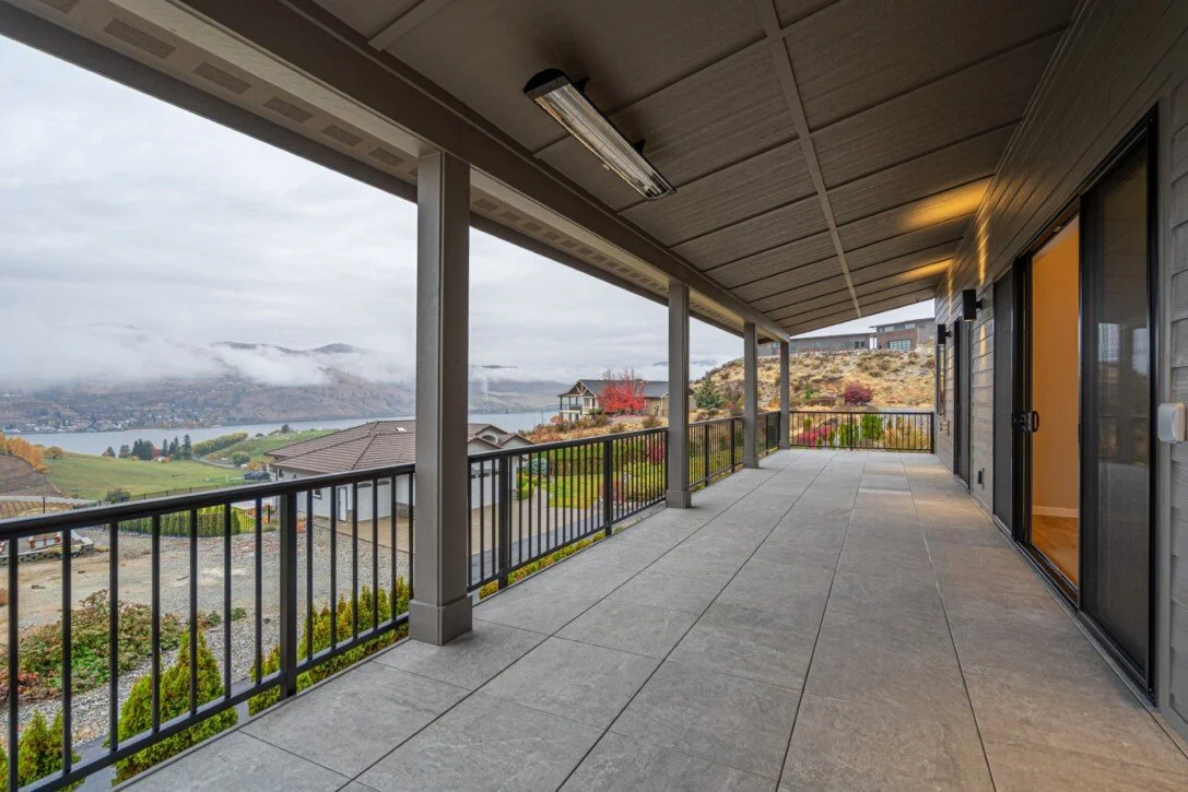 Large covered balcony with grey tiled floor, black railing, and sliding glass doors, overlooking a scenic view of a lake, hills, and houses, with cloudy sky.