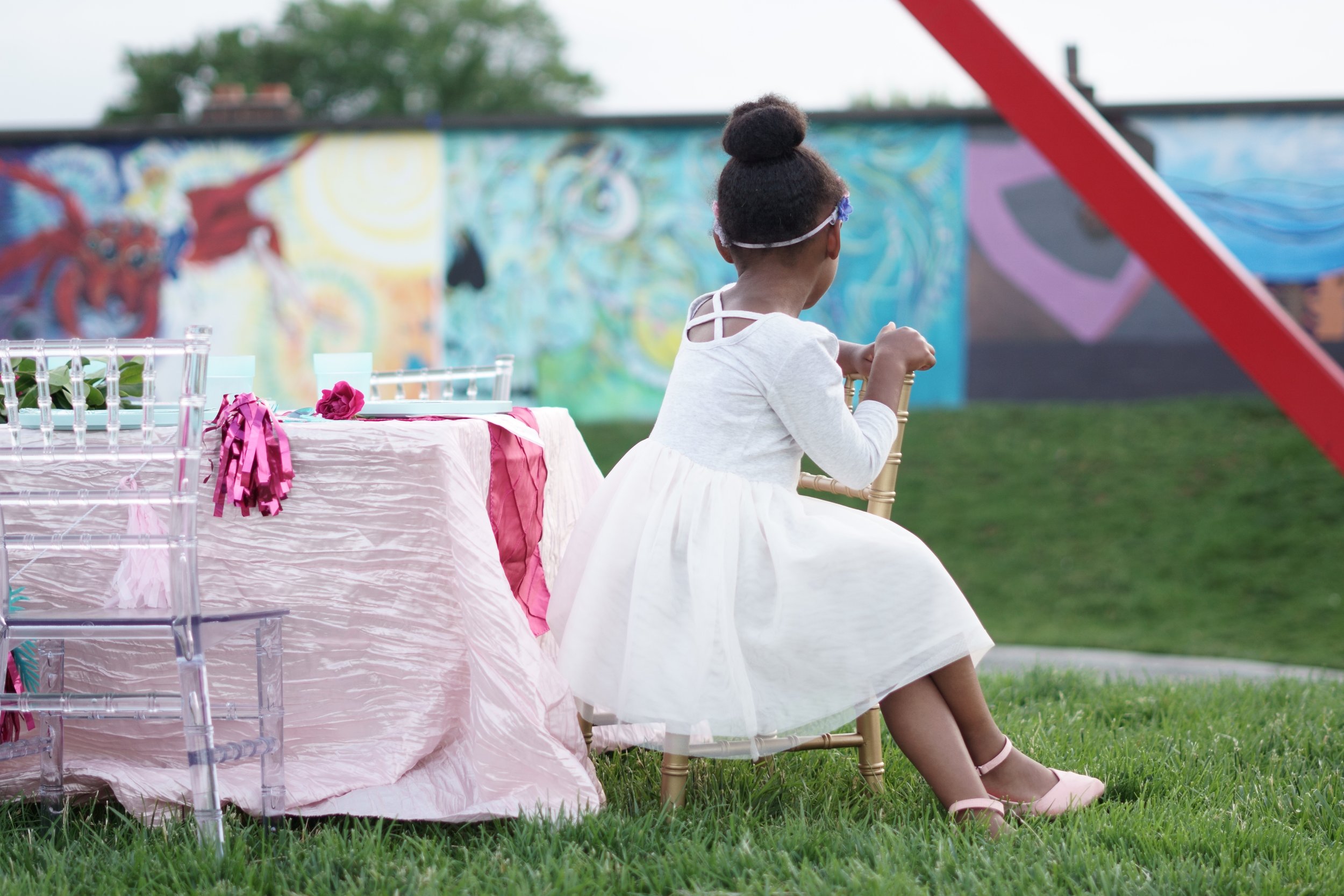 A young girl with dark curly hair tied in a bun, wearing glasses and a white dress, sitting on a wooden chair outdoors near a table covered with a pink tablecloth, set for a celebration with flowers and ribbons. In the background, there is a colorful mural on a wall and a grass field.