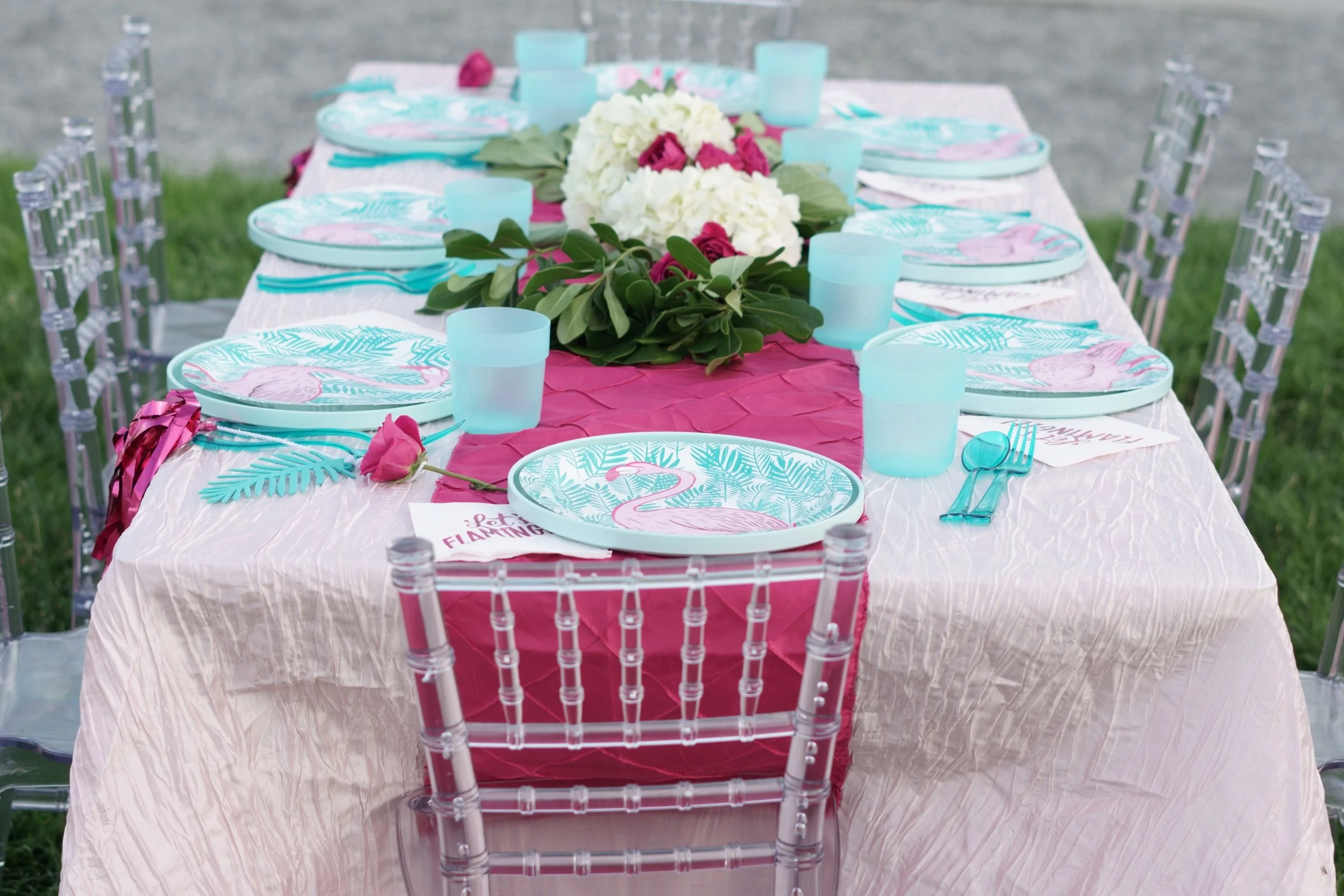 A long outdoor table with a pink tablecloth, set with flamingo-themed paper plates, turquoise cups, and turquoise utensils. The center of the table features a floral arrangement with white and pink flowers and green leaves. Clear chairs are positioned around the table, and the setting appears to be on a grassy area.