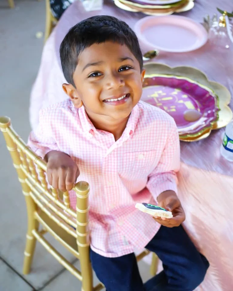 A young boy smiling at a birthday party, sitting on a gold-colored chair at a table decorated for a celebration.