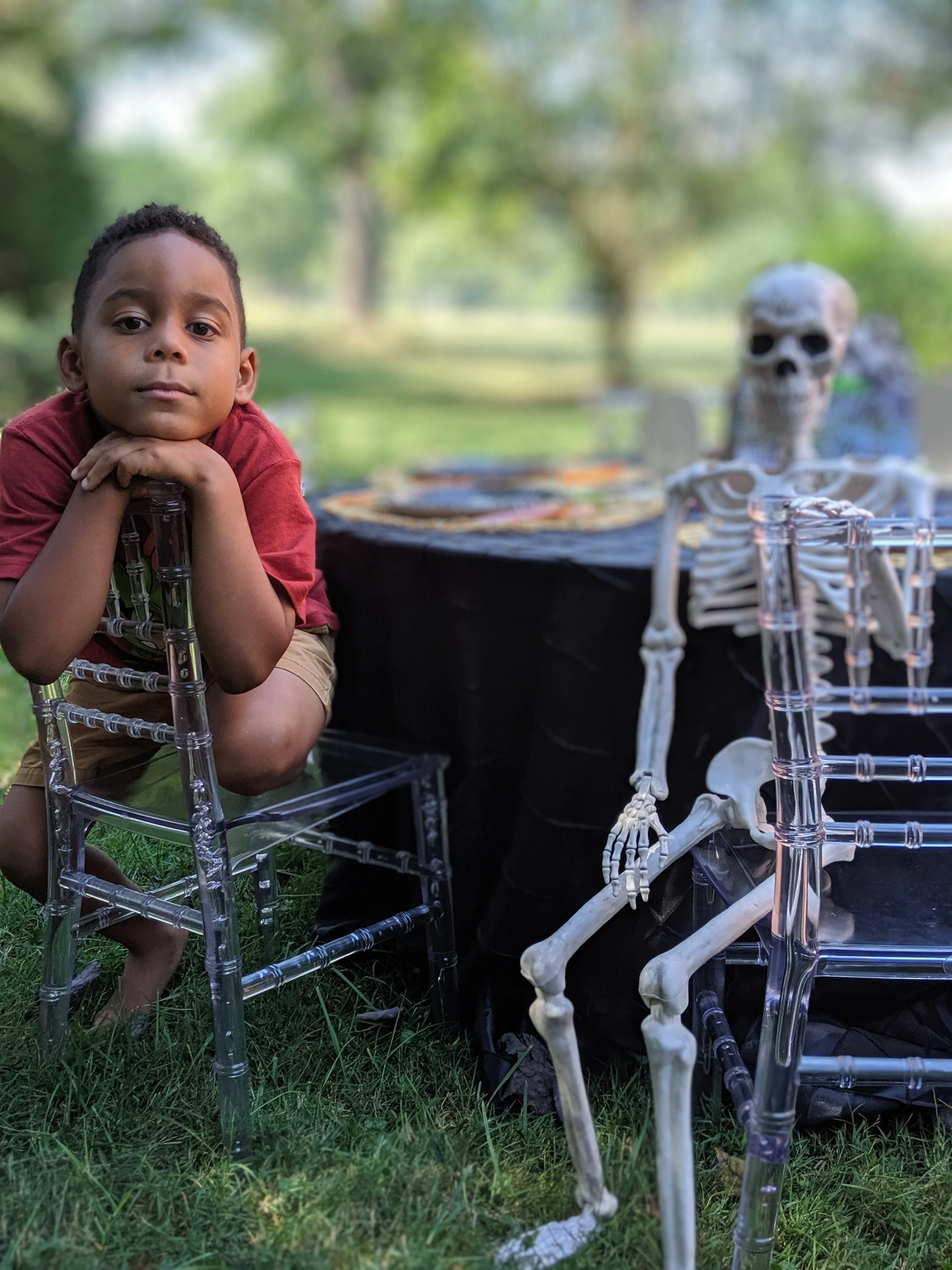 A young boy with short dark hair and a red shirt is sitting outdoors with his chin resting on his folded hands. Next to him is a life-size plastic skeleton and some transparent chairs, set up in a grassy area with trees in the background.