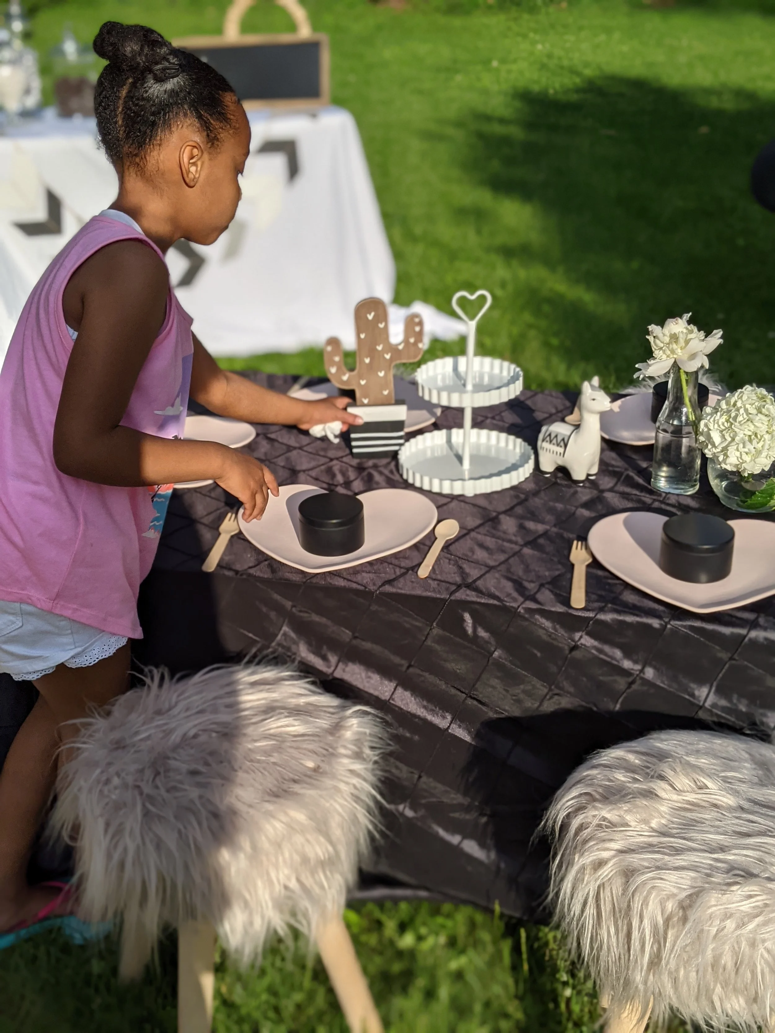 A young girl at an outdoor table with decorative items including a cactus-shaped sign, a white horse figurine, a floral arrangement, and black containers. Her hair is styled in a bun, and she is wearing a pink sleeveless top. The scene is set on a lawn with chairs and tables in the background.