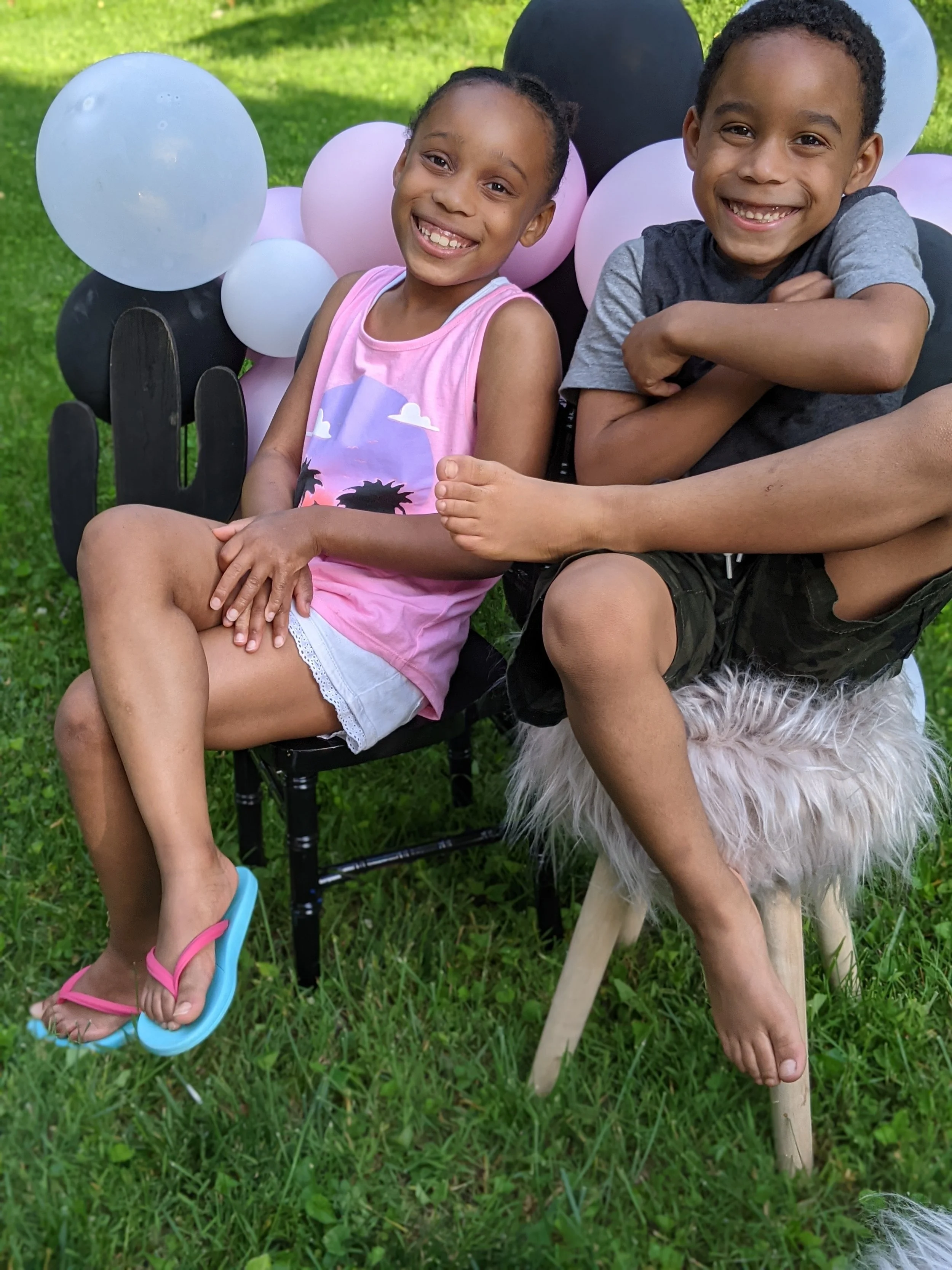 Two children, a girl and a boy, sitting on a black chair outdoors on grass, smiling at the camera with balloons behind them. A dog on a leash is sitting in front of them, partially visible.