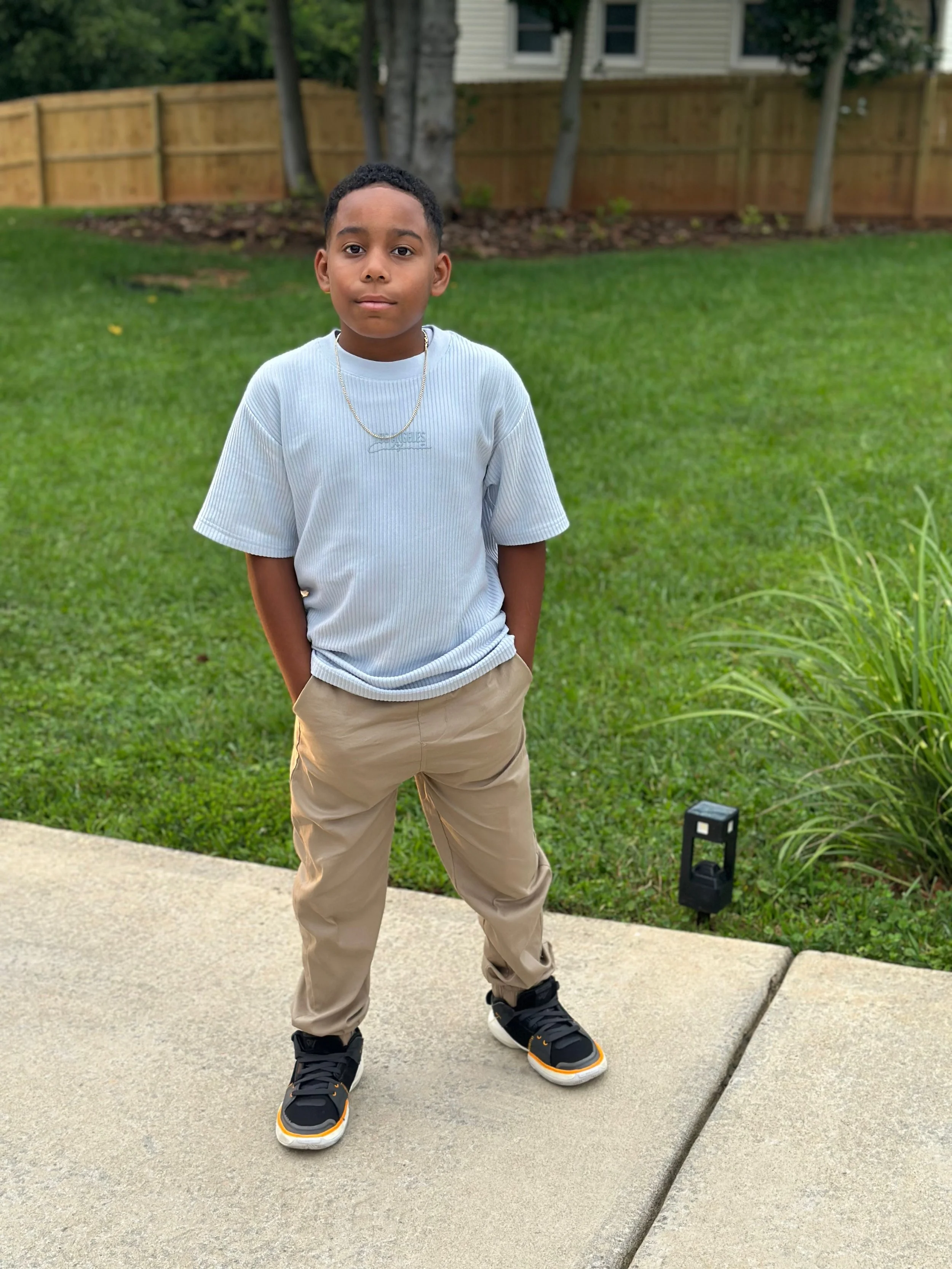 Young boy standing outdoors on a sidewalk with green grass, a wooden fence, and trees in the background, wearing a gray t-shirt, khaki pants, and black sneakers.