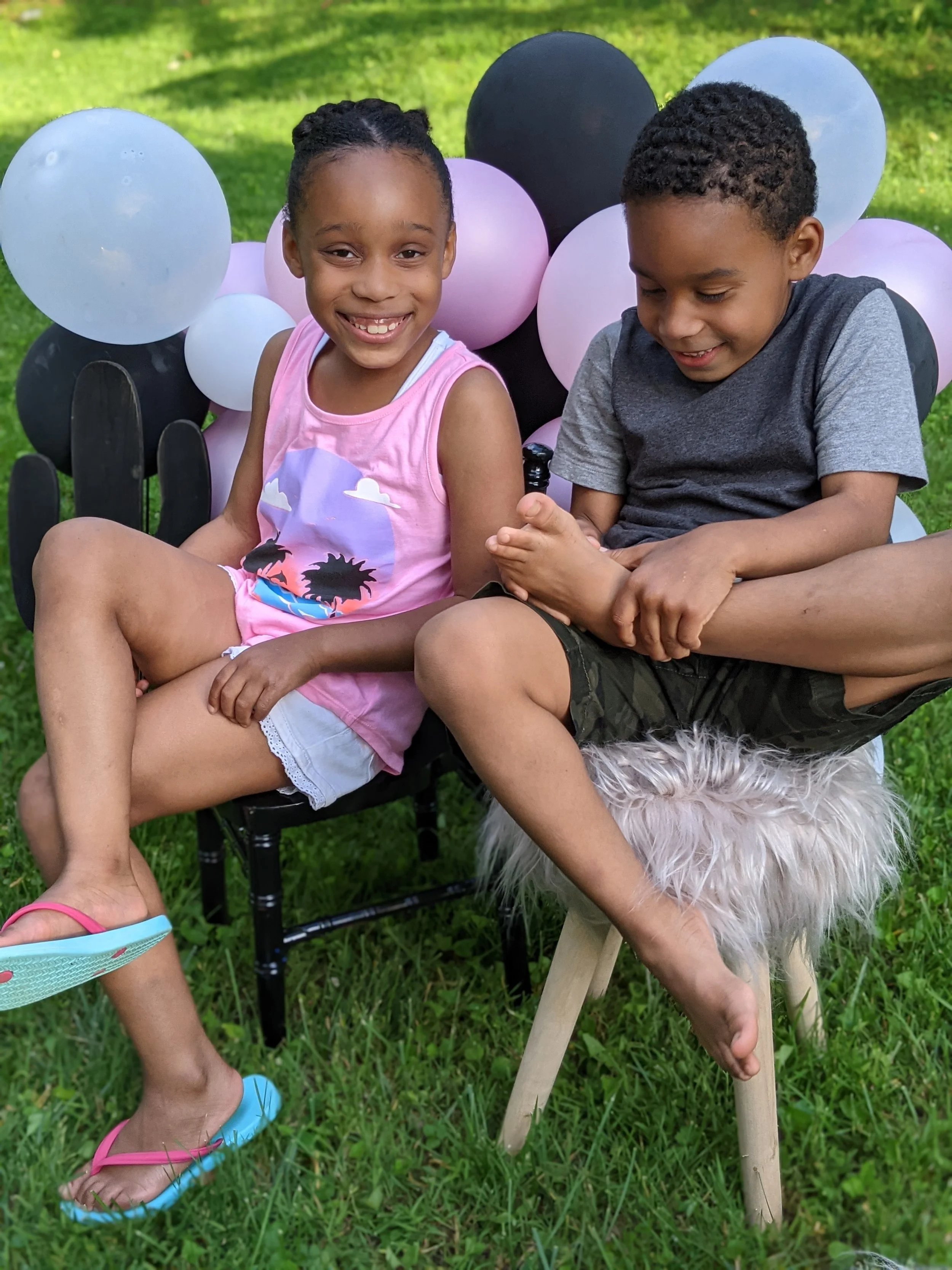 Two kids, a girl and a boy, sitting outdoors on a grassy area, smiling and playing. The girl is wearing a pink tank top with a cloud and sun design, and the boy is wearing a gray T-shirt and camouflage shorts. They are surrounded by colorful balloons and a fluffy stool or blanket underneath the boy's arm.