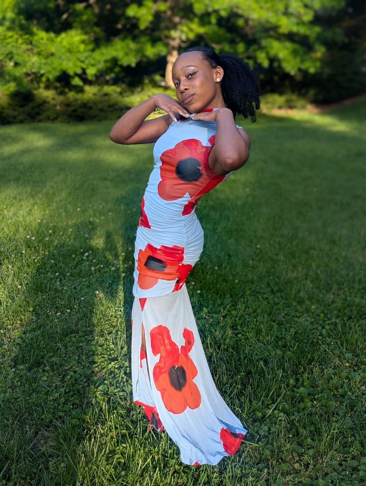 A young woman in a form-fitting white dress with large red and orange poppy flower patterns stands outdoors on grass with green trees in the background. She strikes a pose with her hands near her neck, looking confident.