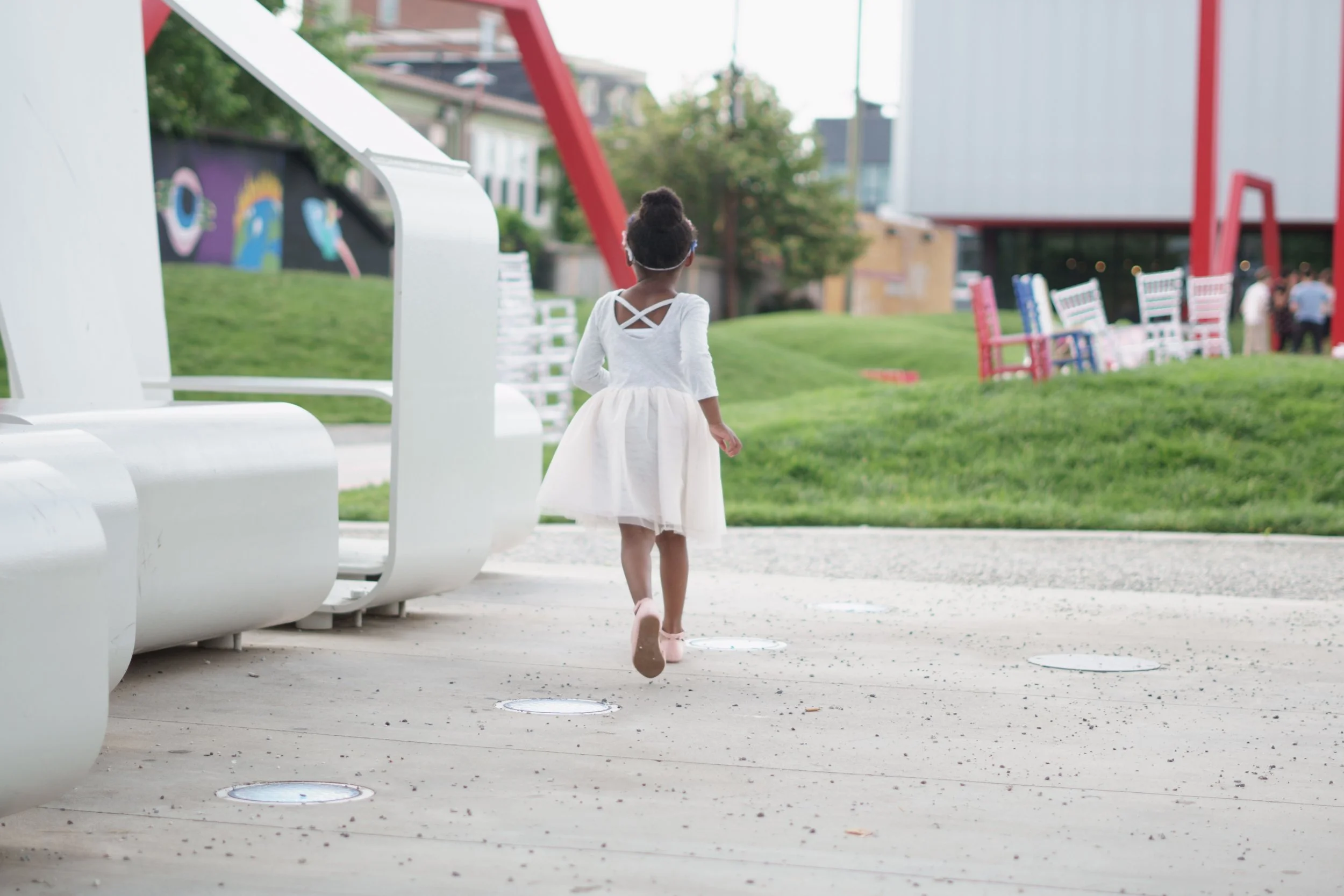 A young girl wearing a white dress with crisscross straps on the back, walking on a paved outdoor area with white circular lights embedded in the ground. She has her hair up and is wearing pink shoes. In the background, there are colorful chairs, a grassy hill, trees, and a building.