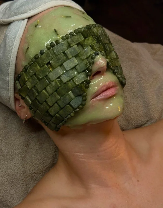 A person receiving a facial treatment with a green mask. The mask is partially covered by a detailed ornamental eye mask made of small green stones and beads, designed to resemble a face for decorative or artistic effect.