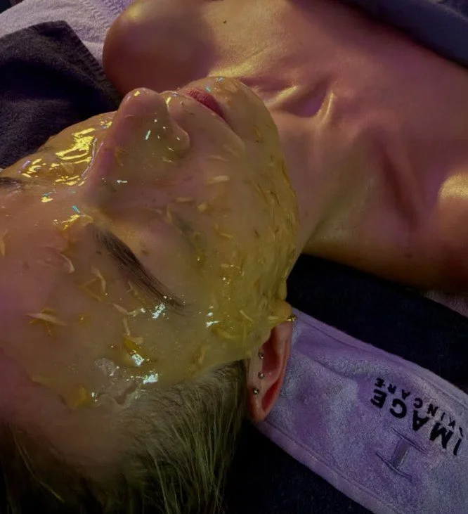 A woman lying down with her face covered in a yellowish, semi-solid substance, possibly a facial mask, and another person lying beside her. The woman has ear piercings and is resting with her eyes closed.