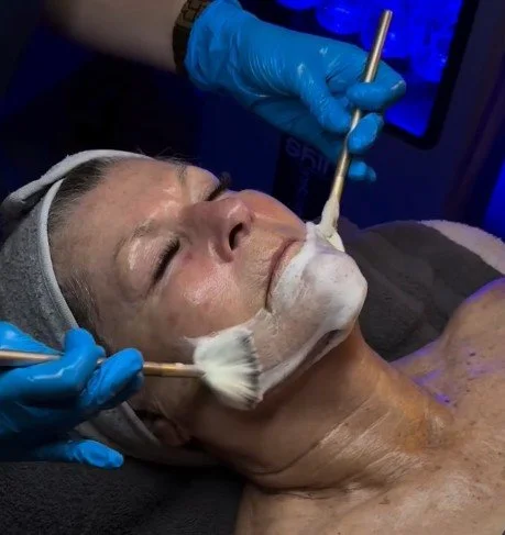 A woman receiving a facial treatment with a brush, under the care of a professional wearing blue gloves.
