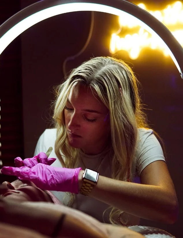 A woman with long blonde hair wearing a white shirt and pink gloves, performing a tattoo on a person's arm under a bright ring light with a warm background.