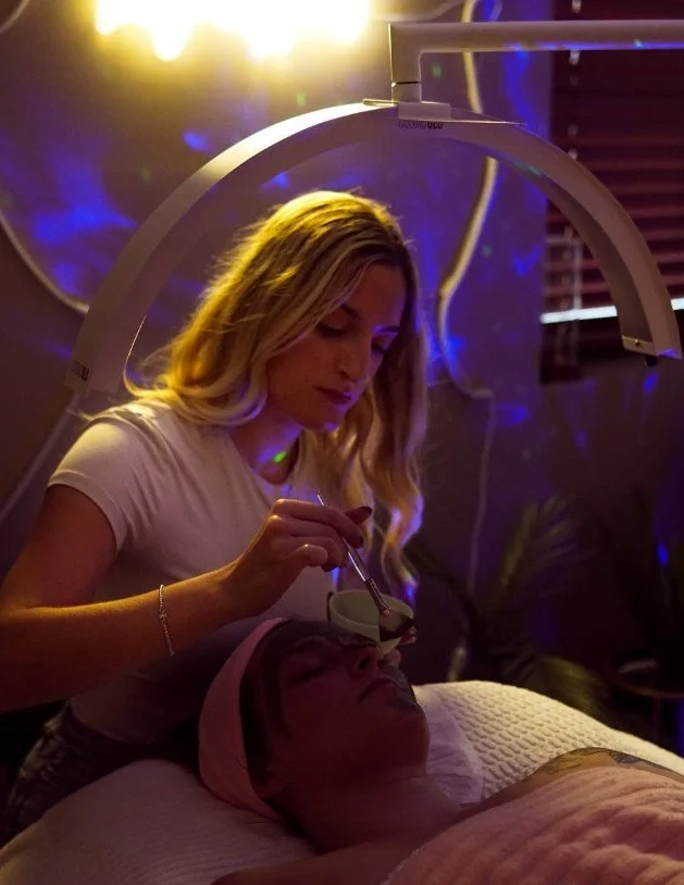 A woman is applying a facial treatment to a person lying in a spa or skincare clinic. The person receiving the treatment has a pink headband, and the setting features warm, ambient lighting with a professional skincare device overhead.