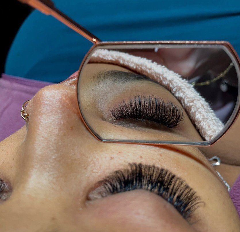 Close-up of a person with eyelash extensions, seen through a mirror. The person is lying down, wearing a headband, with the reflection showing their closed eye with long, thick, curled eyelashes.