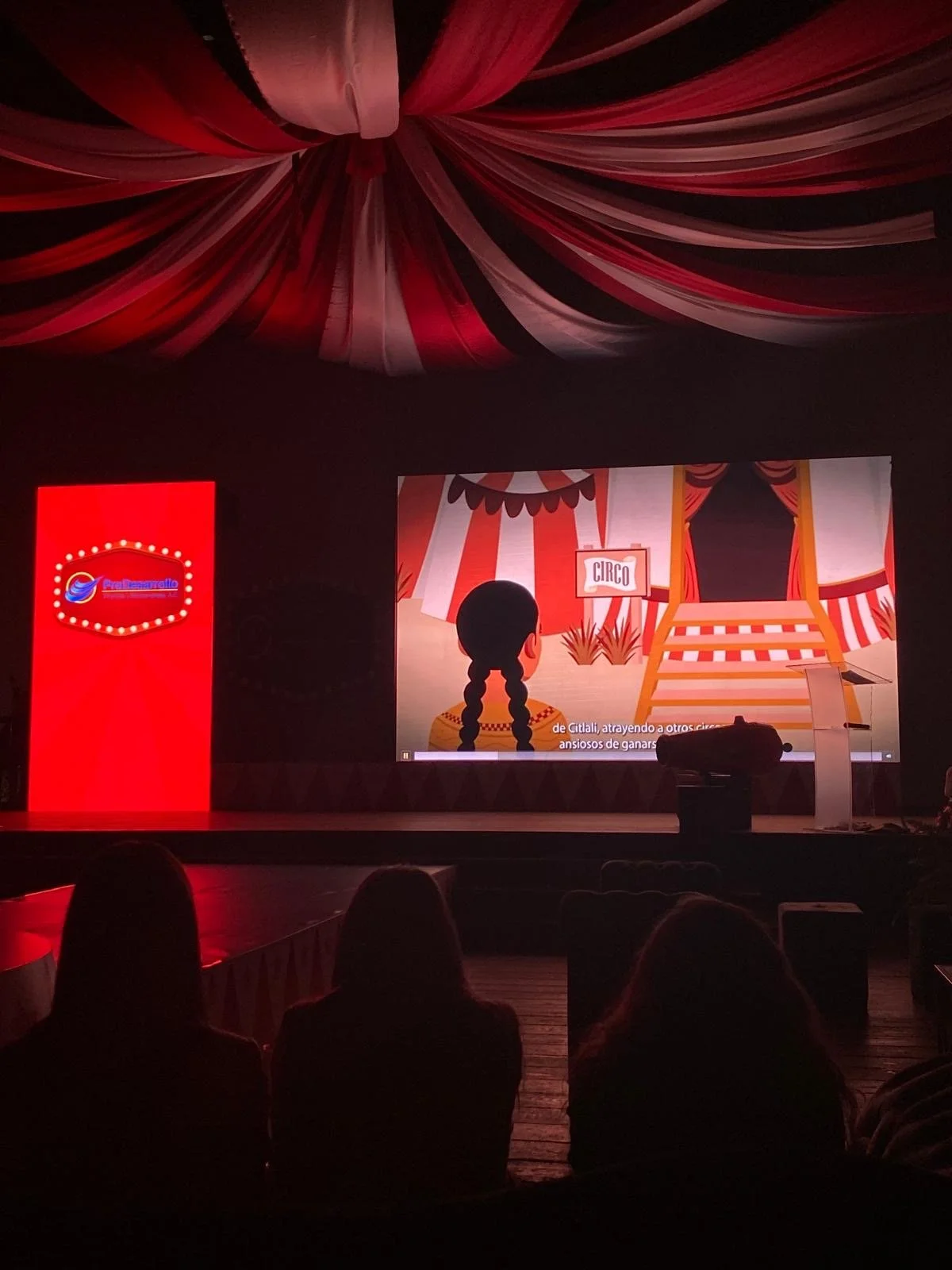 Audience watching a circus-themed animated presentation on a theater stage beneath red and white draped fabric.