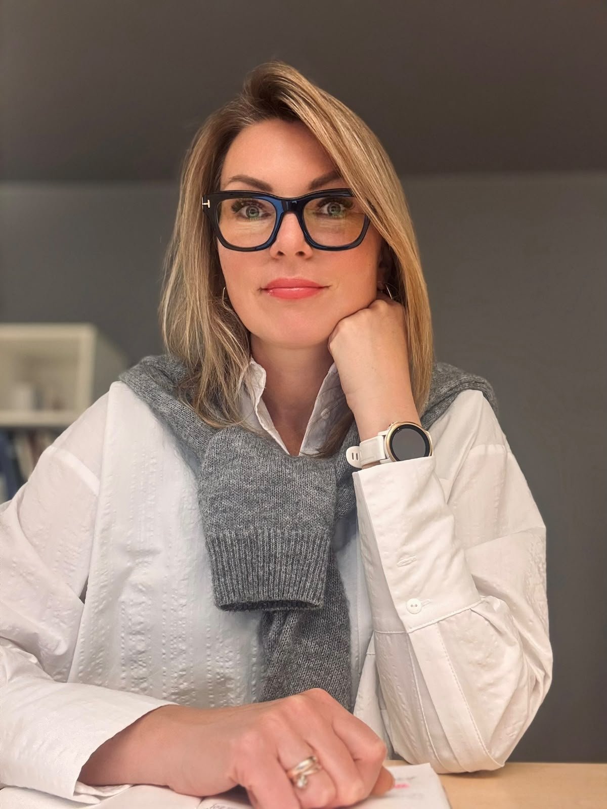 A woman with blonde hair, wearing glasses, a white shirt, and a gray sweater draped over her shoulders, is sitting at a table with her chin resting on her hand, smiling at the camera.