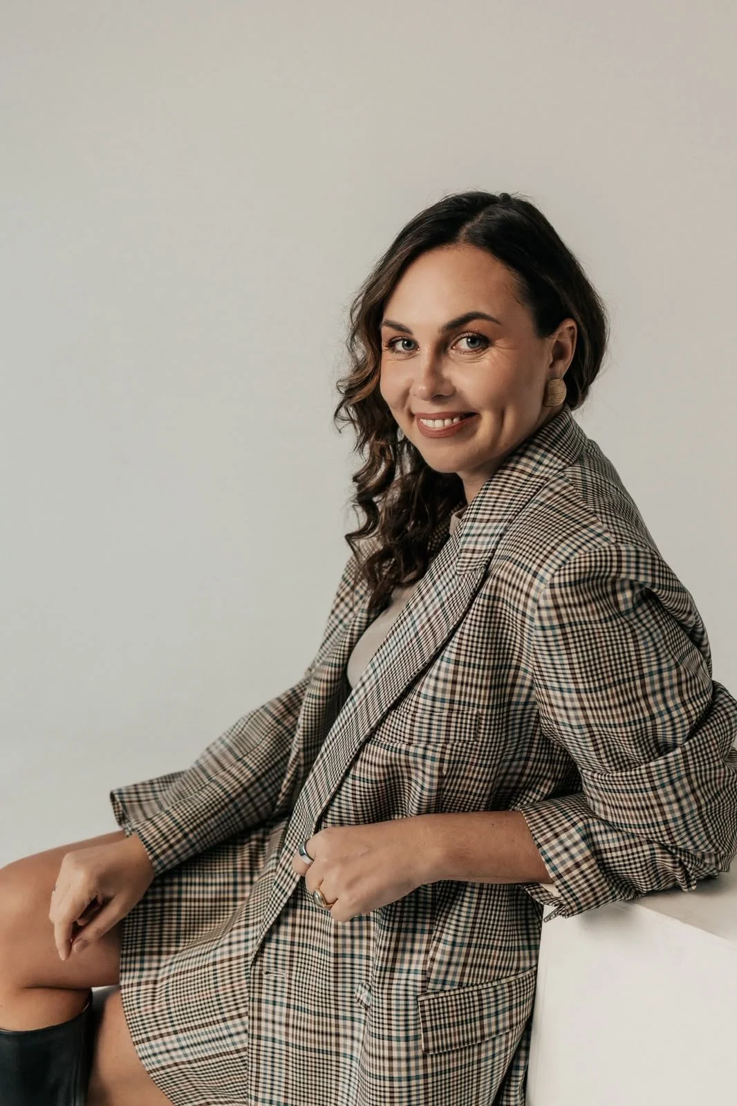A woman with wavy dark hair smiling, wearing a plaid blazer, sitting against a plain light gray background.