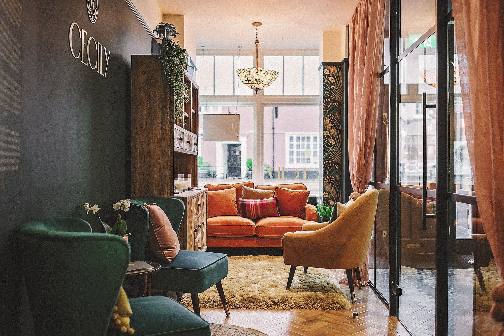 Cozy lounge area with colorful chairs, a wooden cabinet, and a large window letting in natural light, decorated with plants, curtains, and a chandelier.