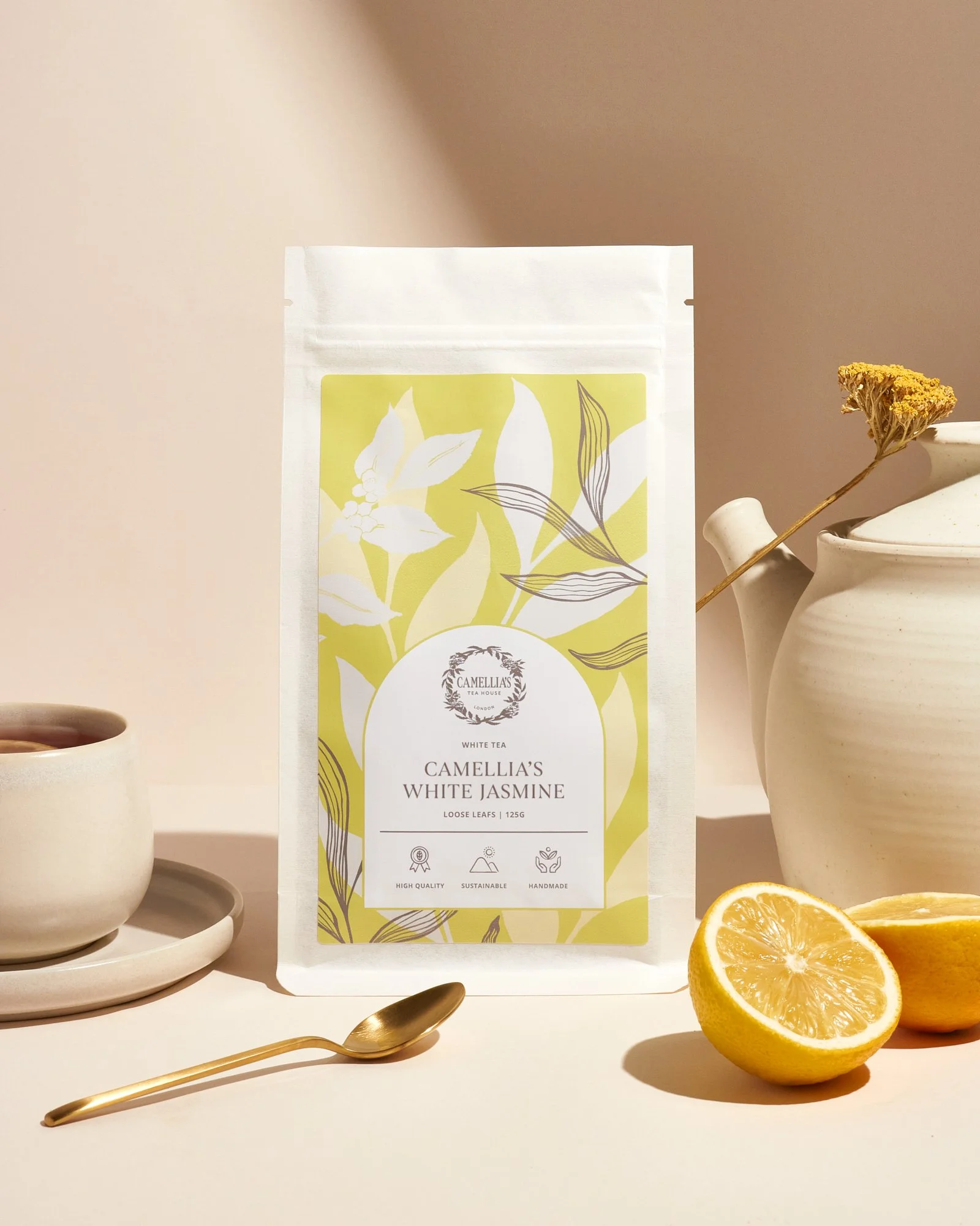 Packaging of Camellia's White Jasmine tea surrounded by white teacup, saucer, golden spoon, halved orange, dried flower, and white vase with a spout, set against a pastel pink background.