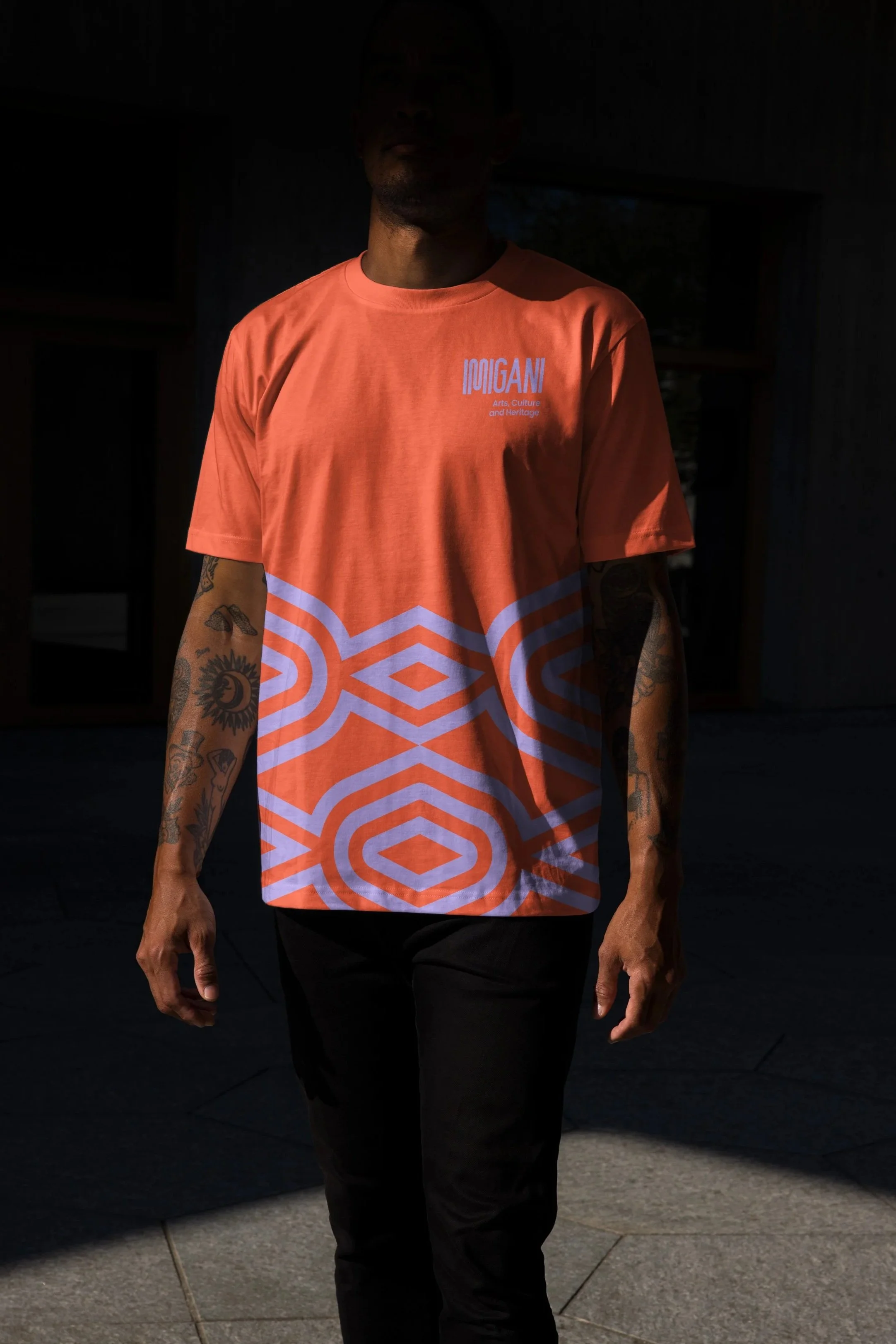 A person with tattoos on their arms stands outdoors in a shadowed area, wearing an orange T-shirt with white text and a geometric pattern.