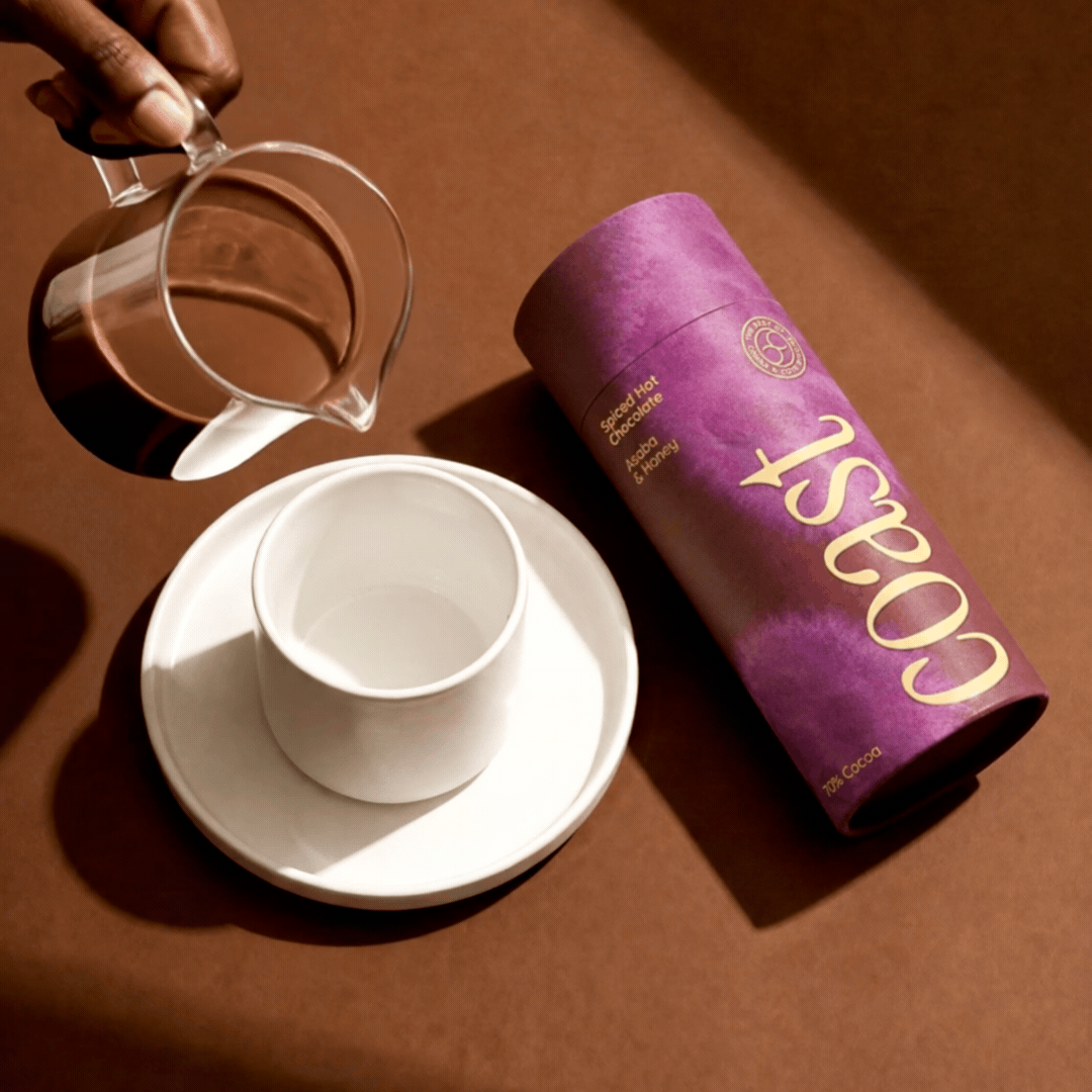 A hand pouring hot chocolate from a clear glass pitcher into a white cup on a saucer, with a purple cylindrical container labeled 'COSY' for spiced hot chocolate with asheo and honey.