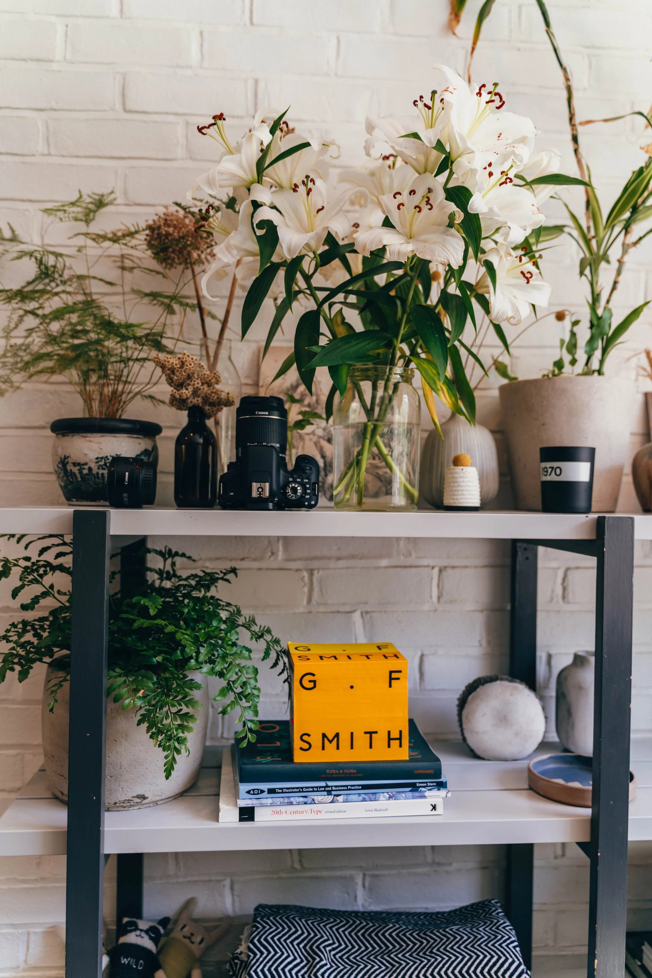 A shelf with a large bouquet of white lilies, various potted plants, a Canon camera and lens, books, and decorative items arranged against a white brick wall.