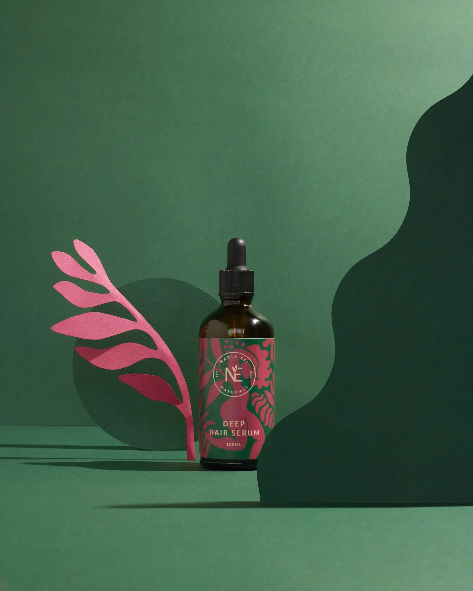 Bottle of deep hair serum with pink and green label featuring a leaf design, placed on green background with decorative pink leaf, dark green abstract shapes.