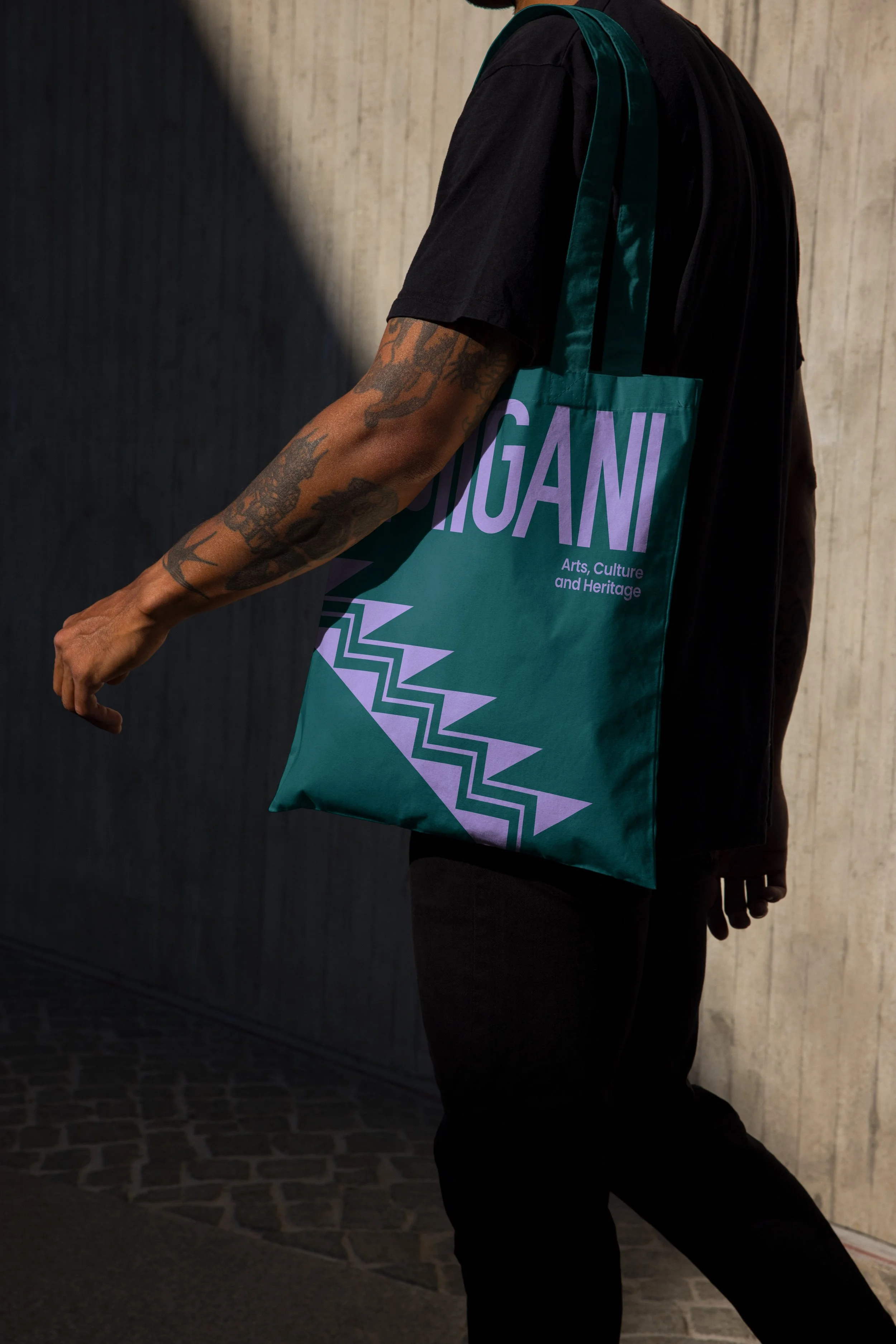 Person wearing black clothes and tattooed arm holding a teal tote bag with purple text that reads "MIGANI" and smaller text that says "Arts, Culture and Heritage." The person is standing against a textured wall, and the image captures their torso and arm.