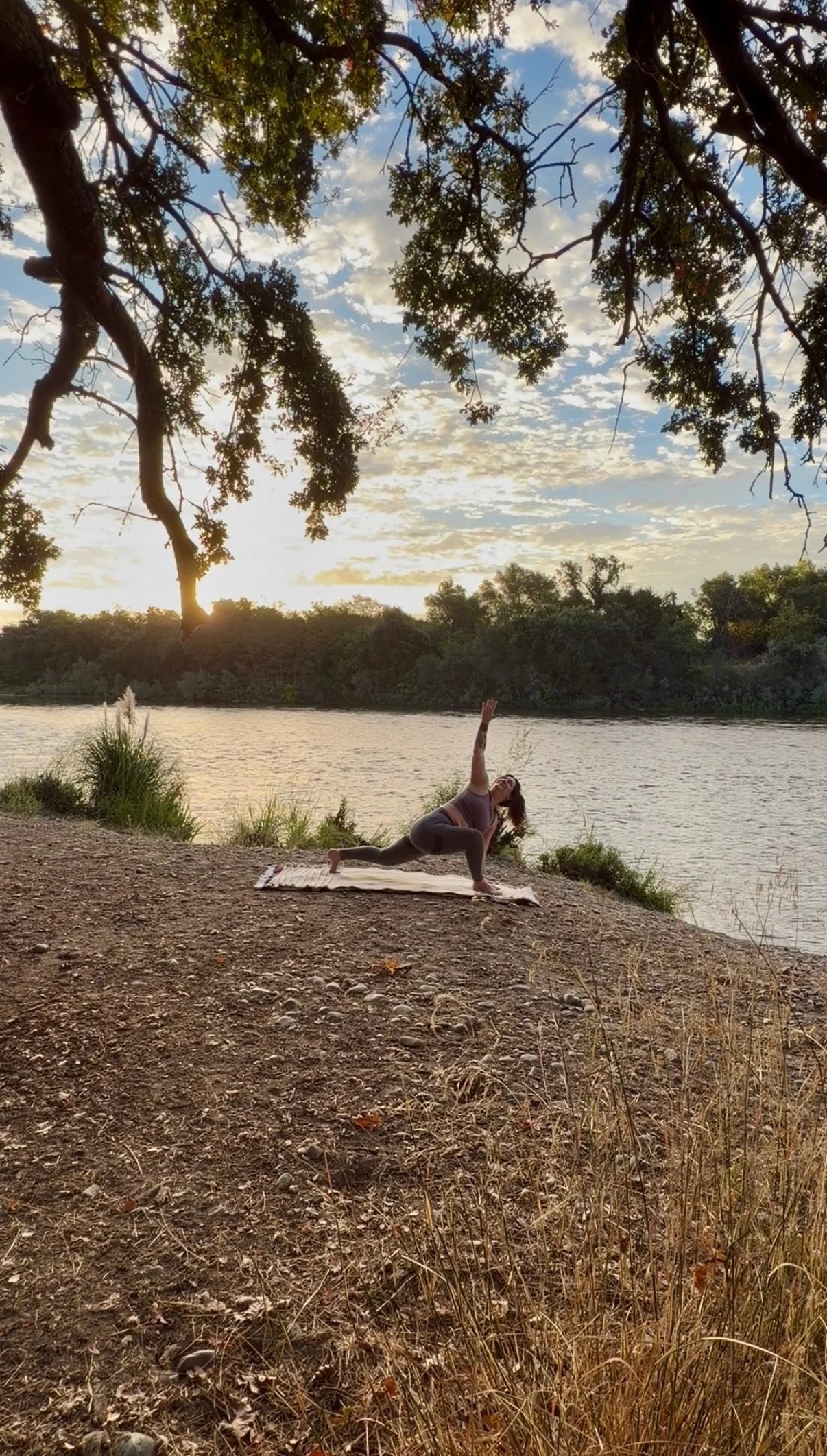 A woman practicing yoga outdoors near a river during sunset, performing a low lunge pose with one arm reaching upward, under a tree with clouds and blue sky in the background.