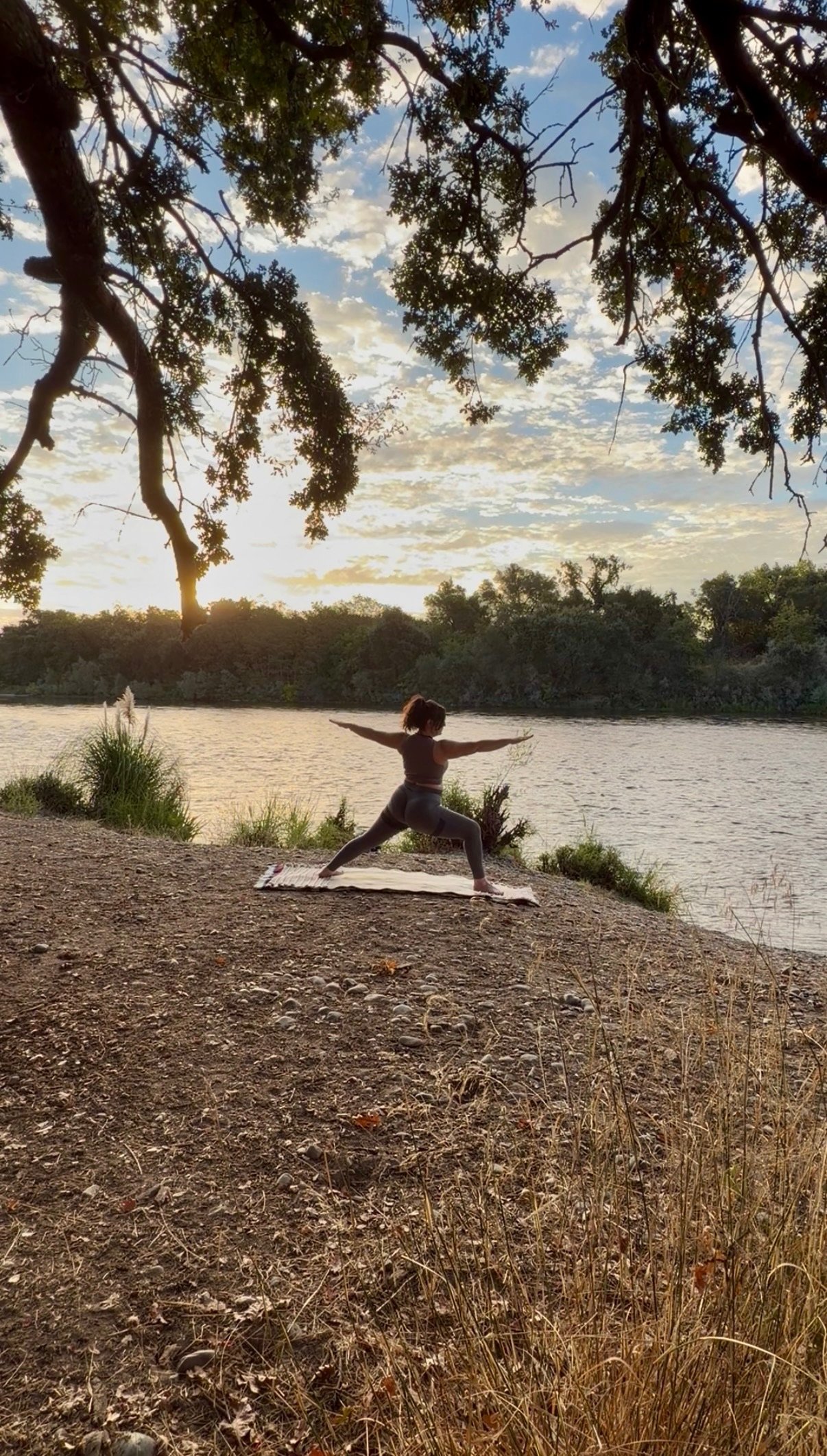 A woman practicing yoga outdoors by a river during sunset, performing Warrior pose on a yoga mat beneath large trees.