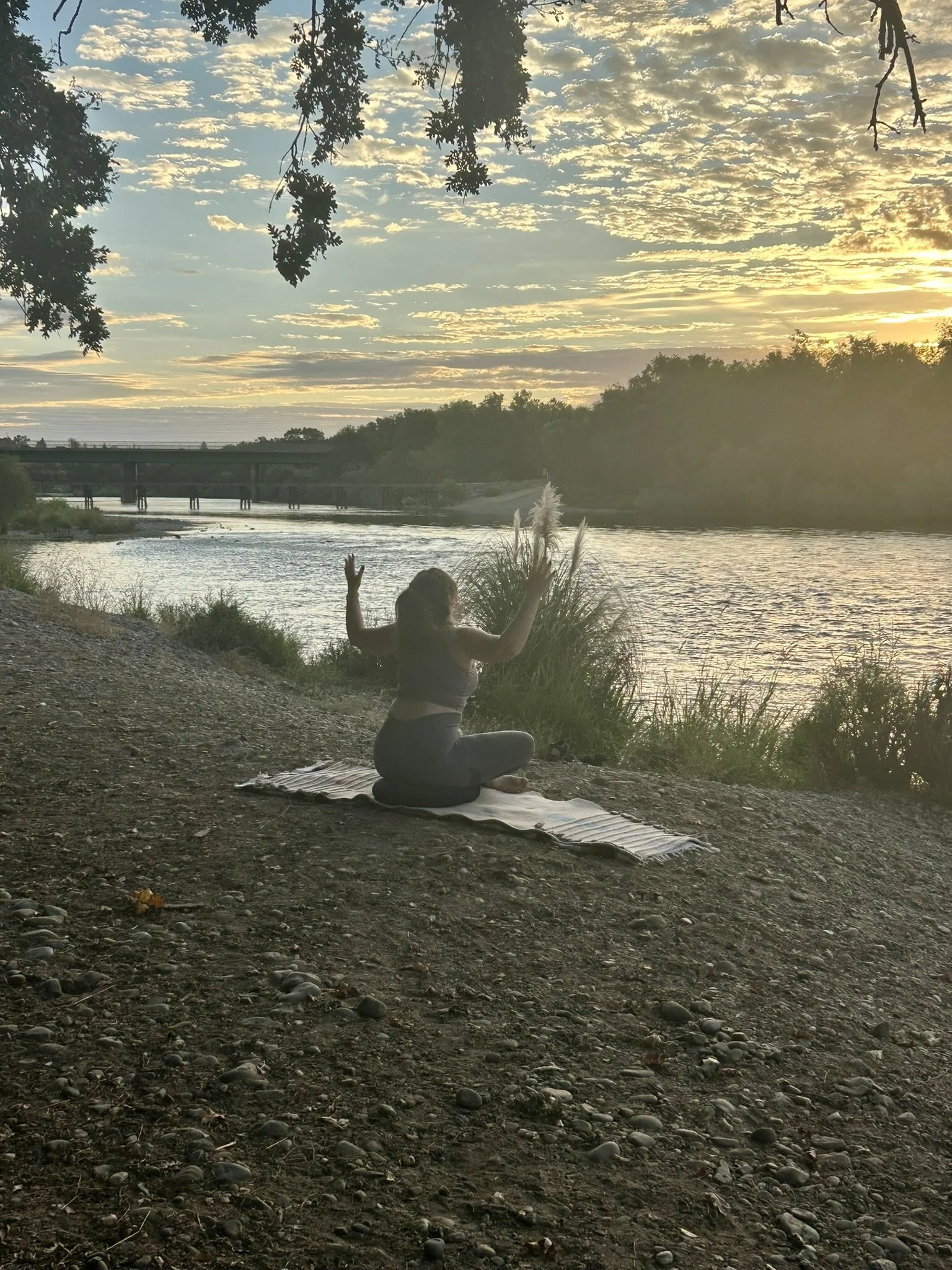 A person practicing yoga in a seated meditation pose on a mat by a river at sunset, with trees and a bridge in the background.