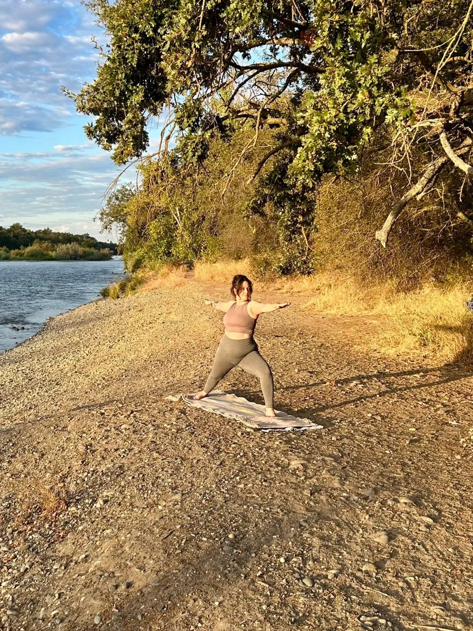 A woman practicing yoga outdoors by a lake, performing Warrior pose on a yoga mat, with trees and a partly cloudy sky in the background during late afternoon or early evening.