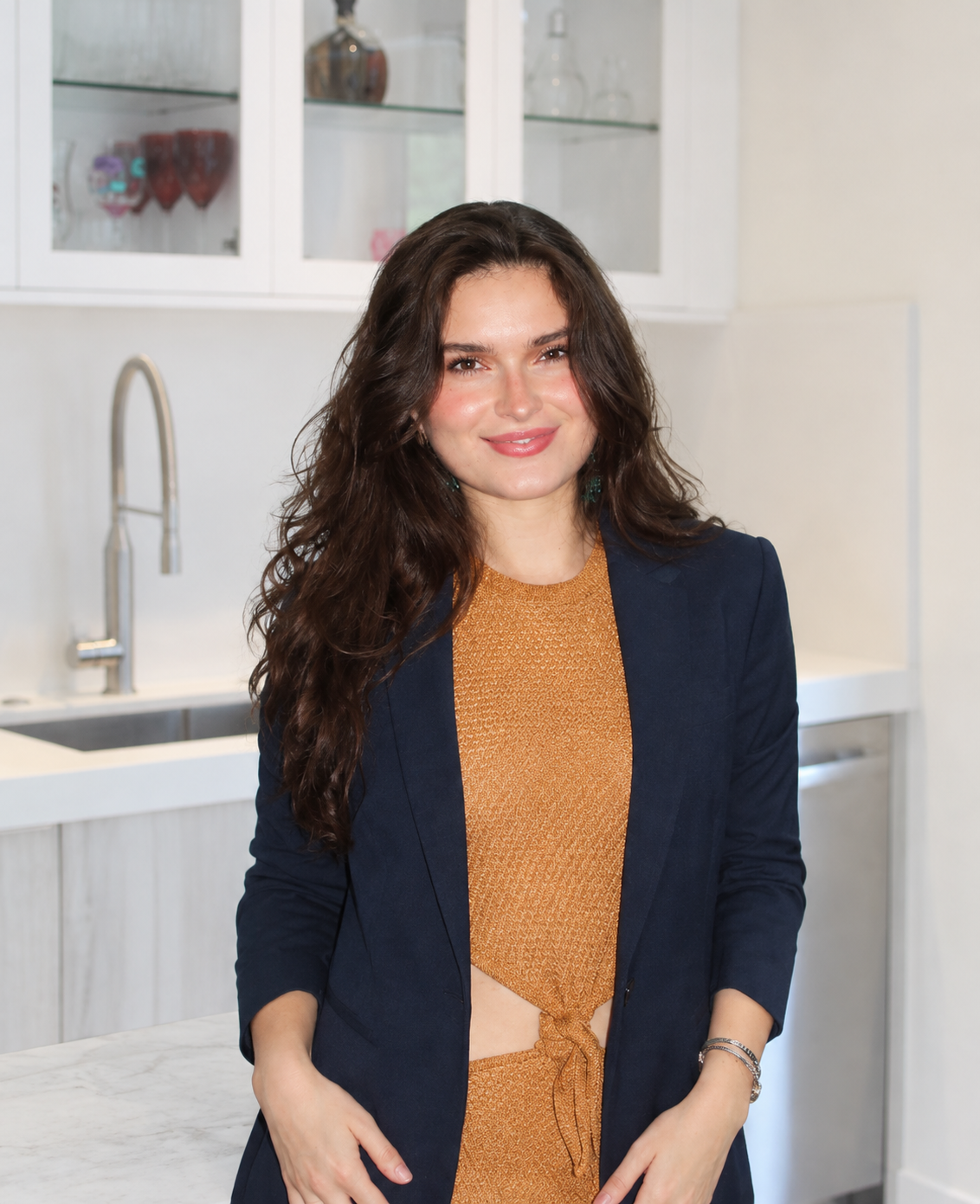 A woman with long, wavy brown hair and a friendly smile, wearing a navy blazer over an orange dress, standing in a bright kitchen with white cabinets and a marble countertop.