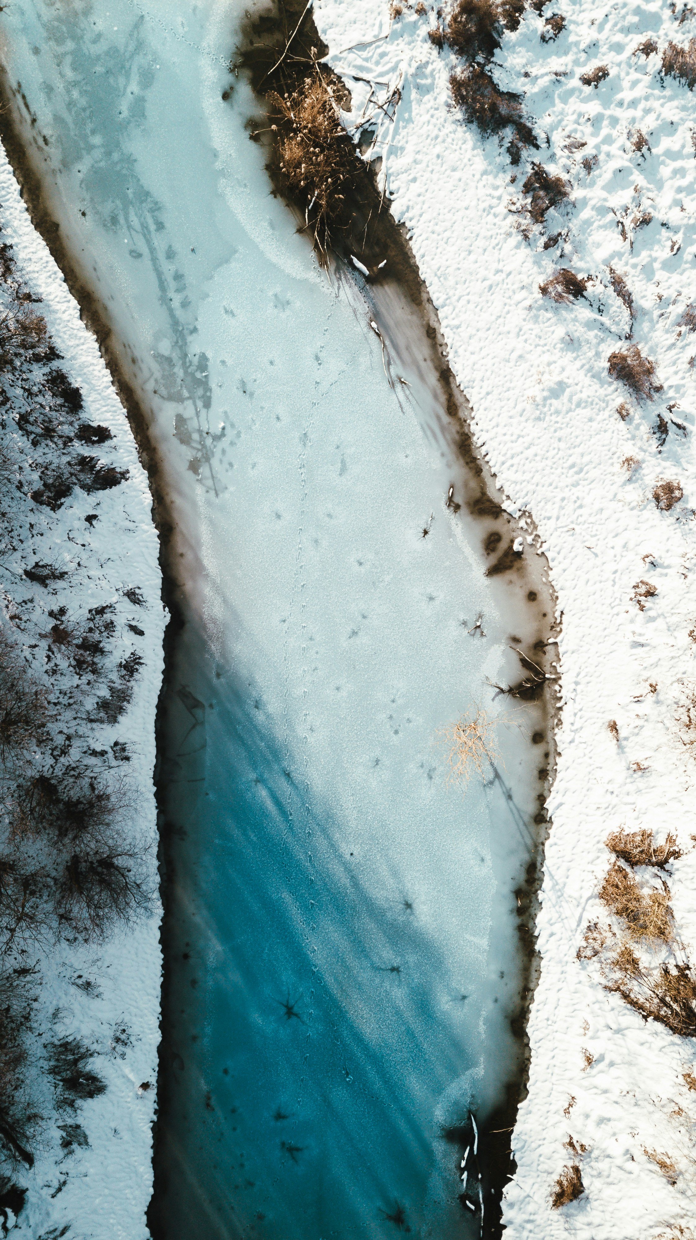 Bird's-eye view of a snow-covered riverbank with a partially frozen river flowing through it, surrounded by snow and sparse trees.