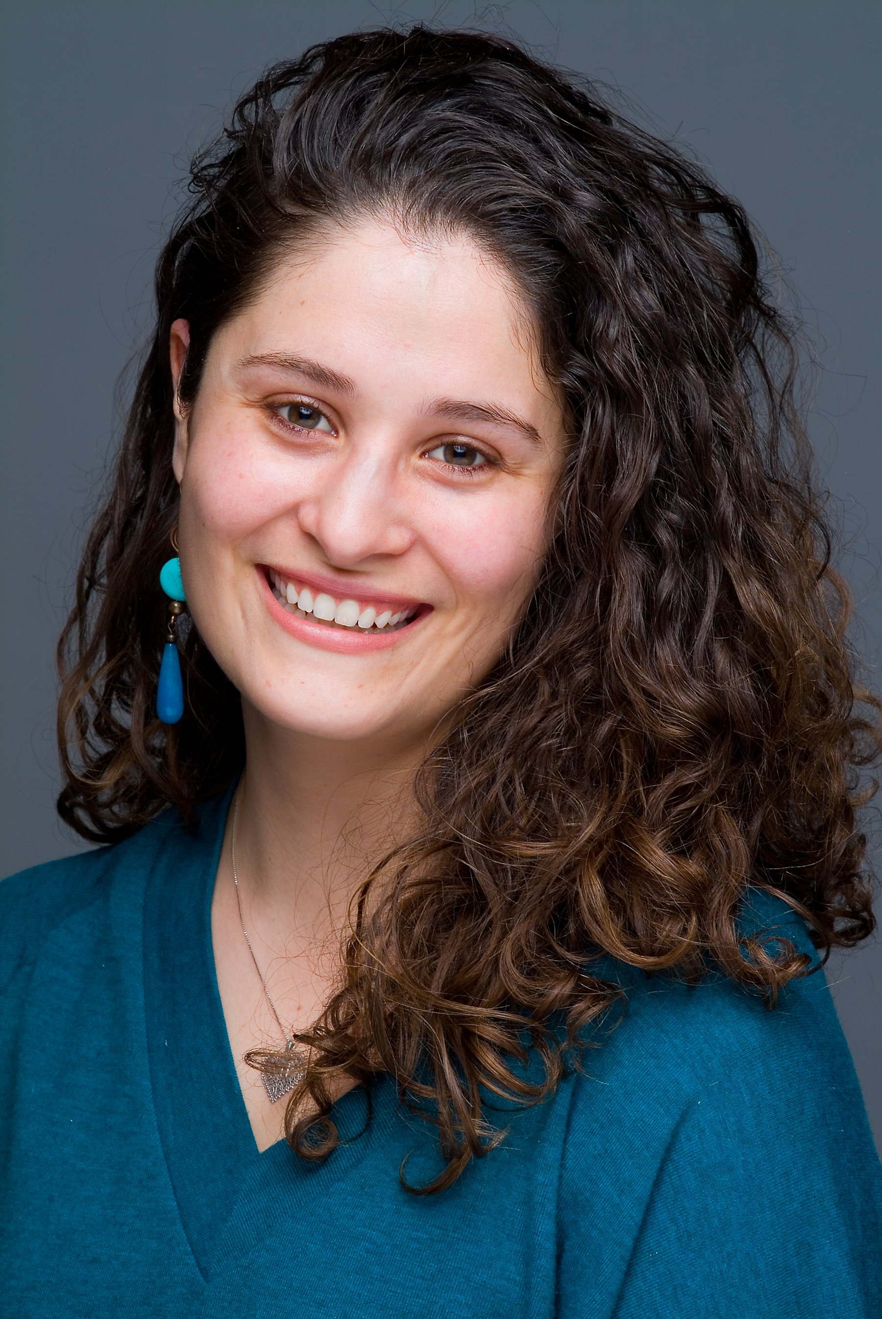 A woman with long, curly brown hair smiling, wearing a teal top, turquoise earrings, and a silver necklace with a butterfly pendant, against a dark gray background.