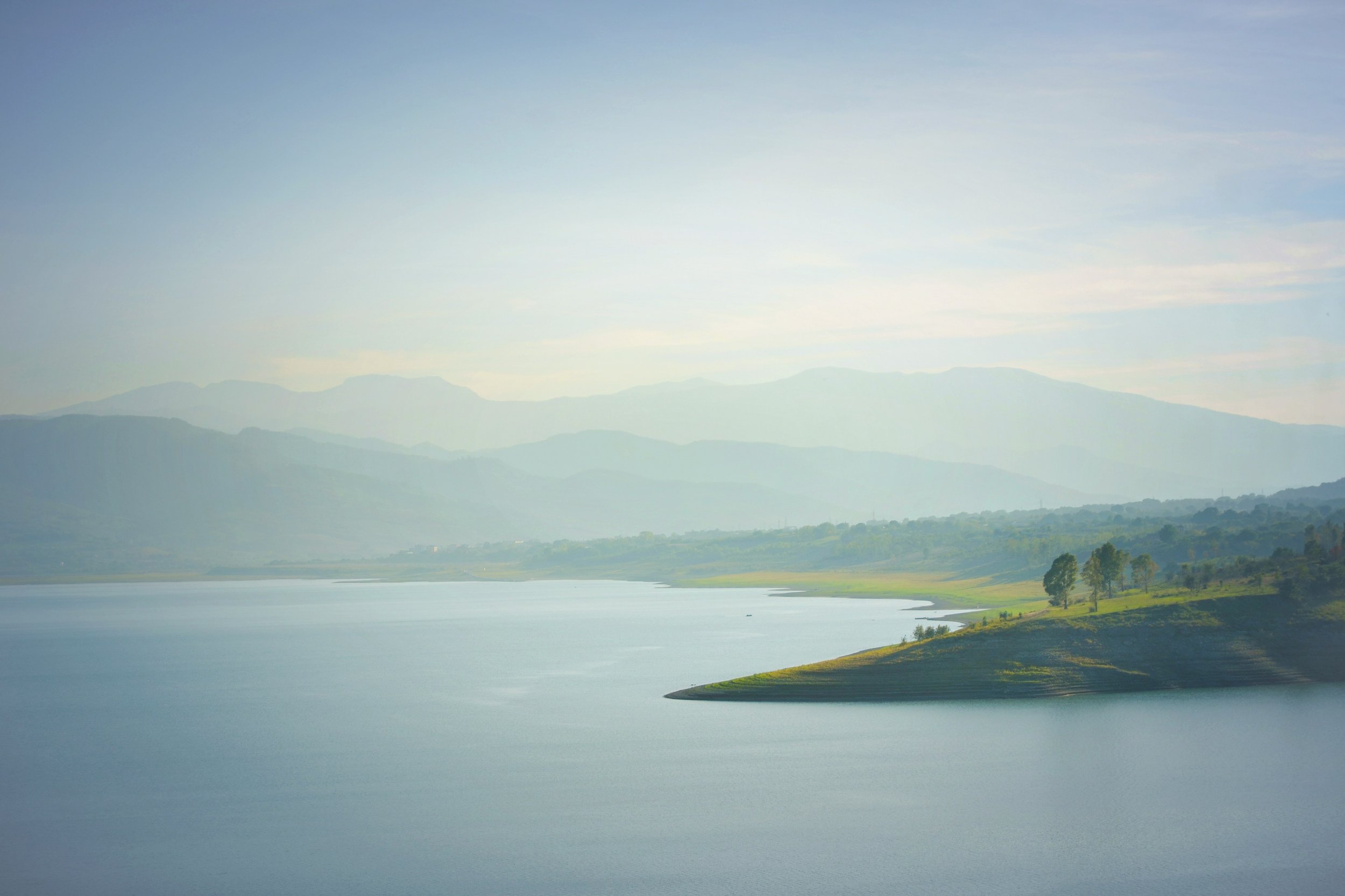 A serene landscape featuring a large body of water with faint reflections, lush green shores, rolling hills, and distant mountains under a clear, pale blue sky.
