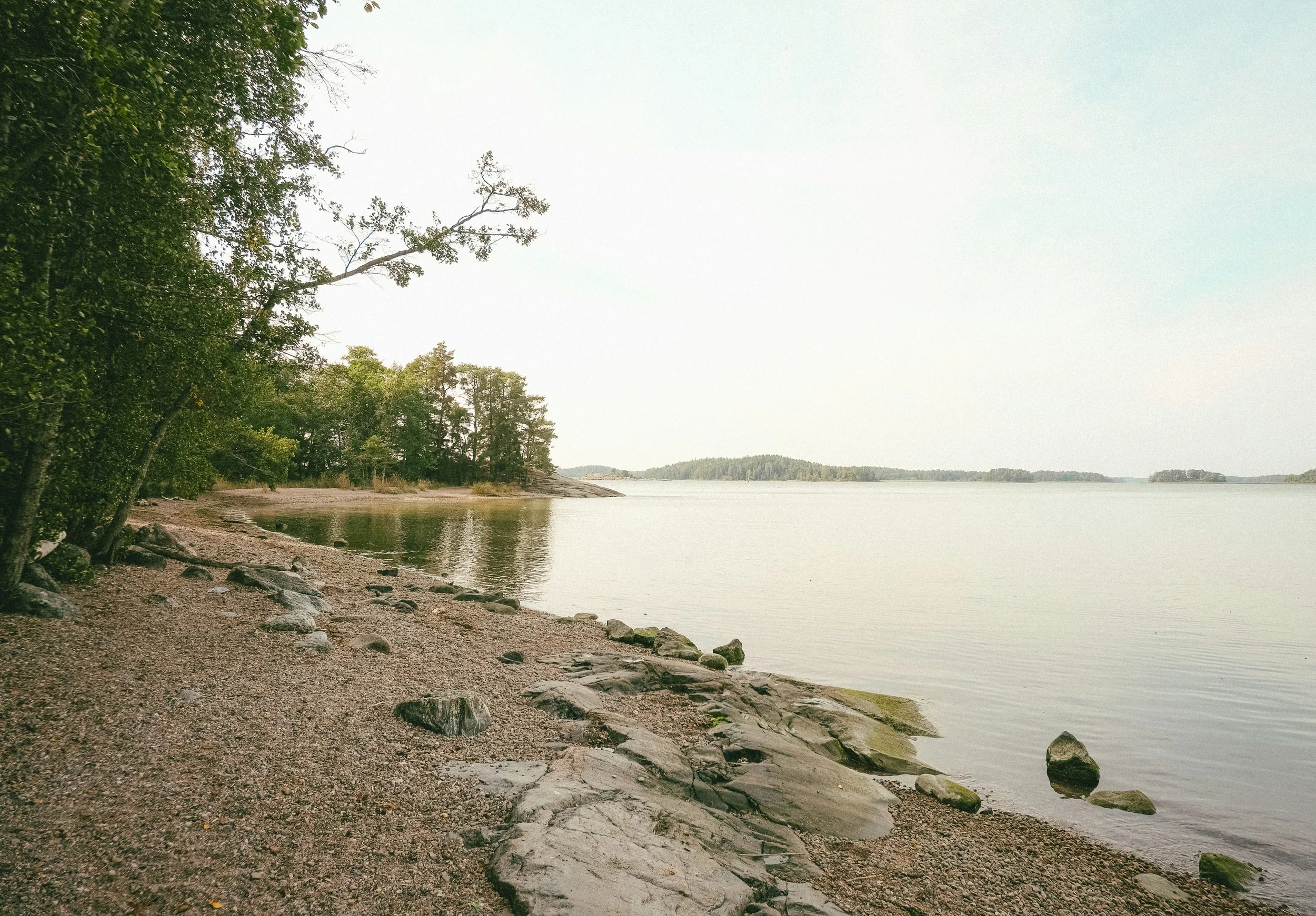A serene lakeside scene with a rocky and sandy shore, green trees on the left side, calm water extending into the distance, and a clear sky overhead.