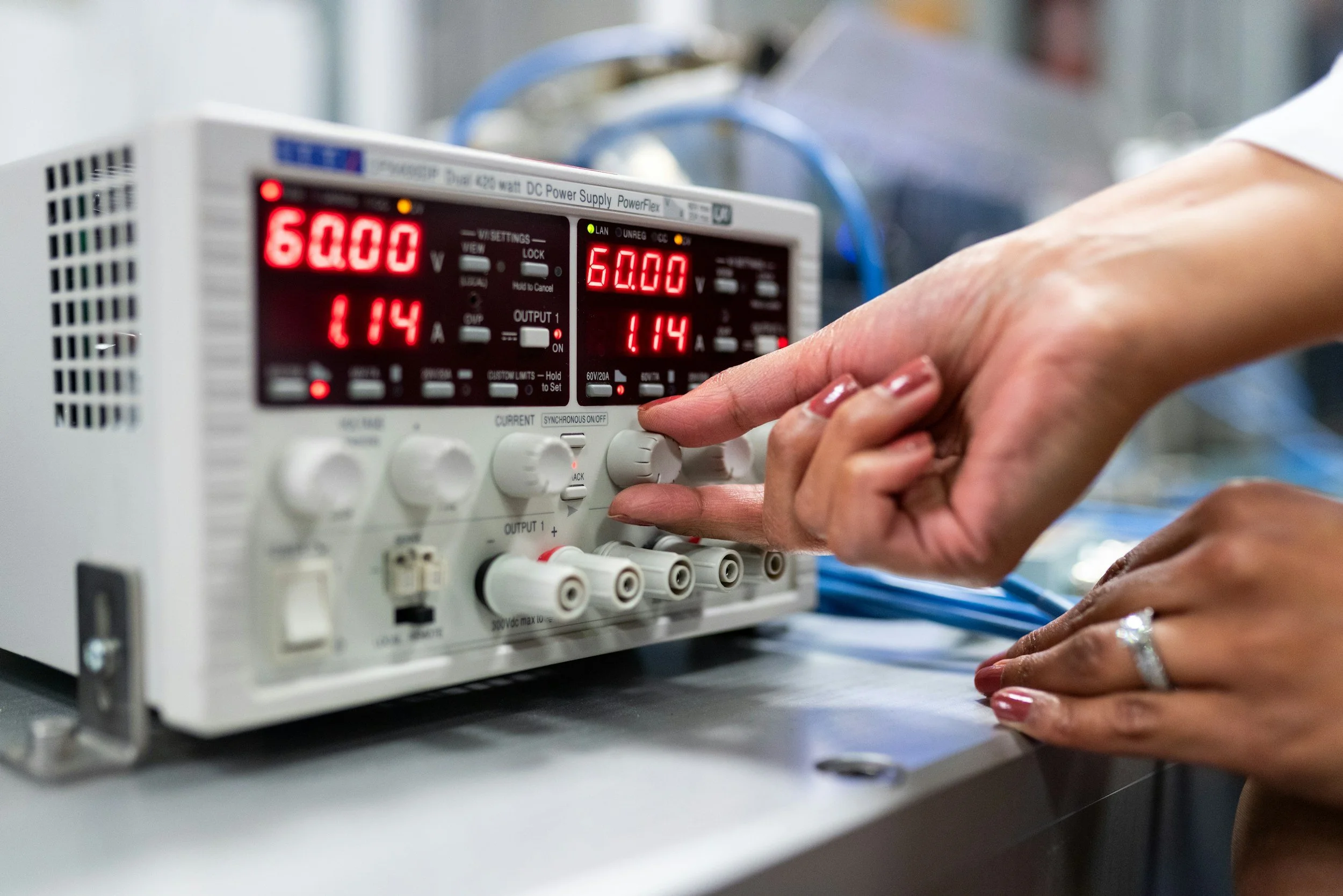 Close-up of a person's hand adjusting a digital power supply device with red and green LED displays, showing voltage and amperage readings.