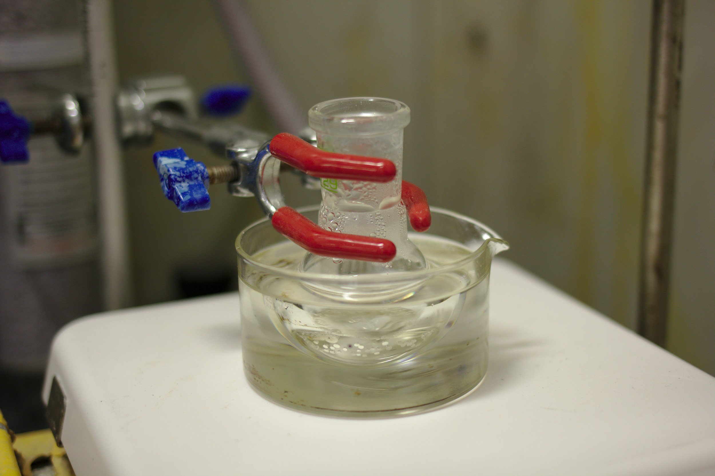 A laboratory setup with a glass container submerged in water, surrounded by red and blue tubing, with a metal clamp holding the tubing in place. The setup is placed on a white surface.