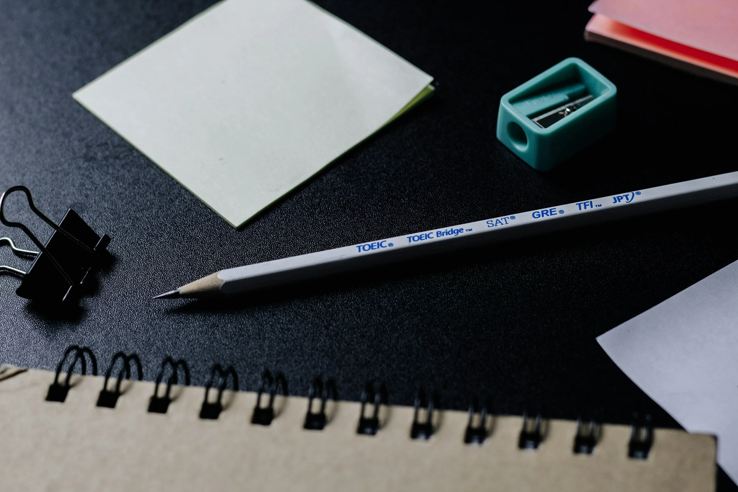 A black textured desk with a spiral notebook, a pack of correction tape, a white pencil, a binder clip, a pile of sticky notes, and an academic test booklet with various standardized test names printed on the white pencil.