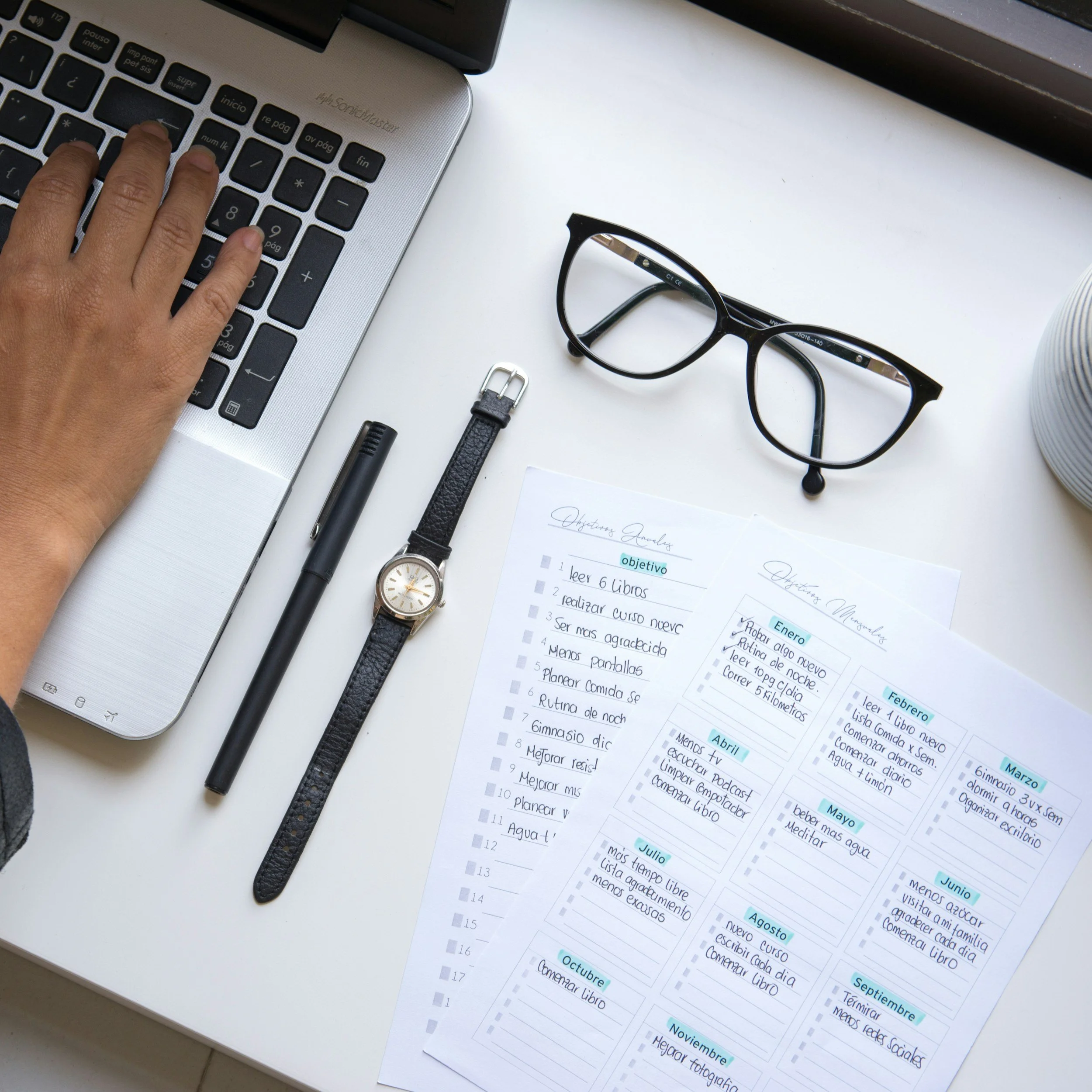 A workspace with a person's hand typing on a laptop keyboard, surrounded by eyeglasses, a wristwatch, a black pen, a paper clip, and printed sheets with handwritten and printed notes in Spanish. A white cup or container is partially visible on the right side.