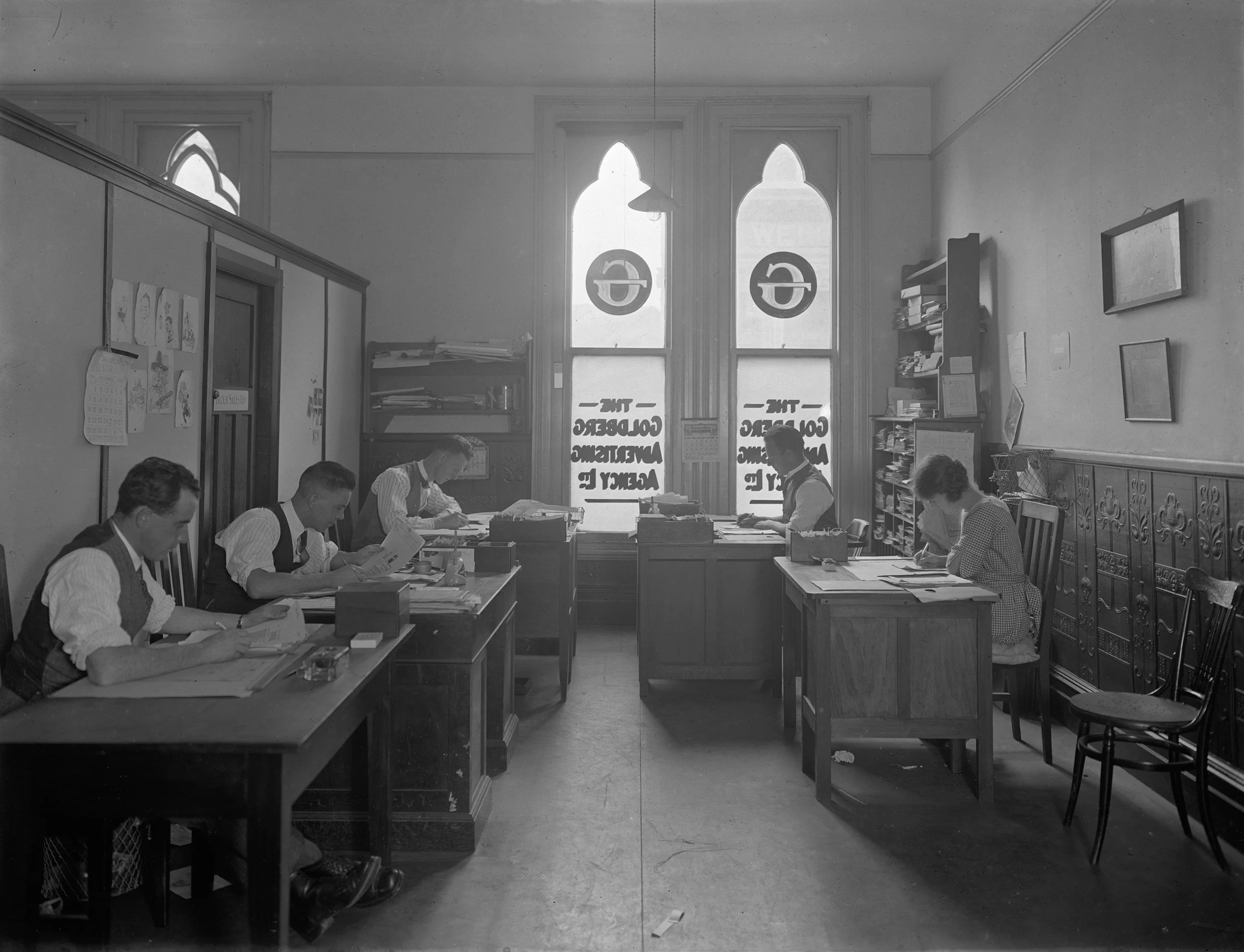 Black and white photo of a classroom with boys and girls sitting at desks, reading or working. Tall windows with Gothic arches and circles at the top are in the background. Shelves with books and papers, framed pictures, and a bulletin board are on the walls.