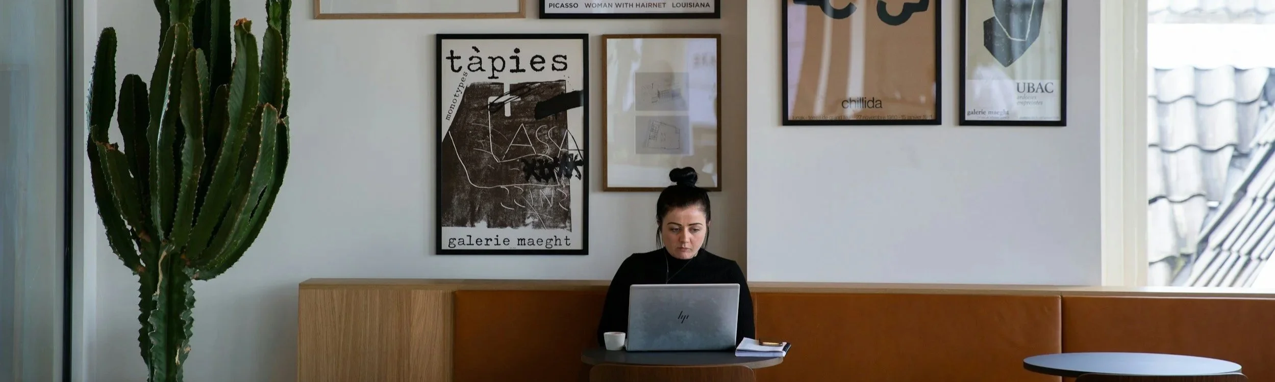 A woman sitting at a wooden table working on a laptop in a modern, art-filled office space with a cactus plant to the left and framed posters on the wall behind her.