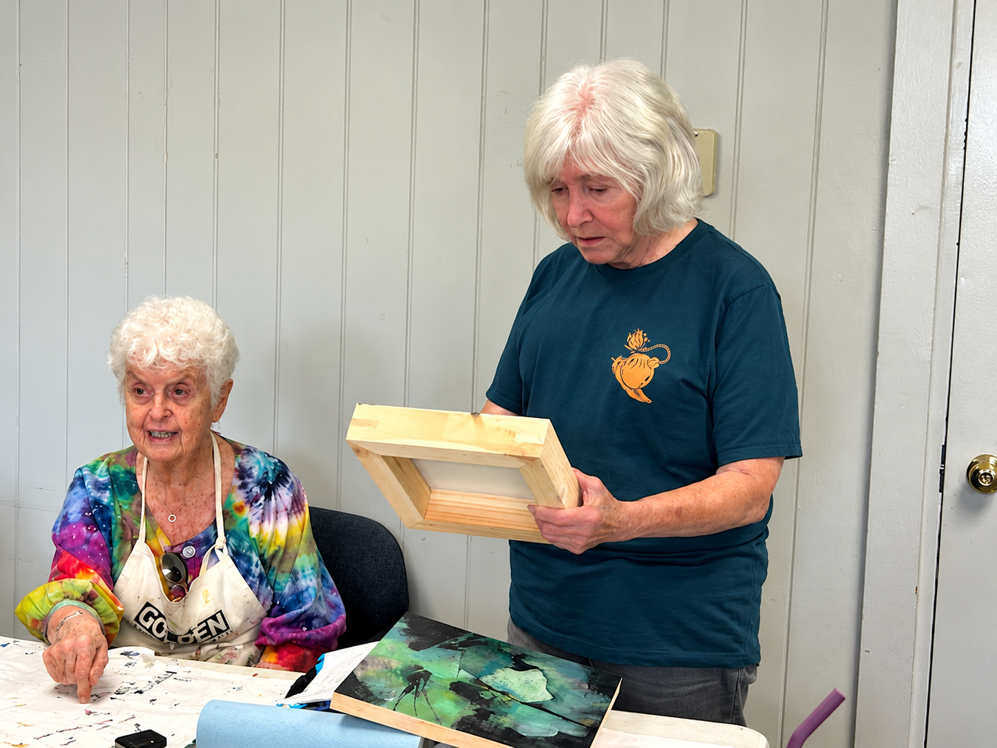 Two older women at an art table, with one holding a wooden frame and the other wearing a paint-splattered apron.
