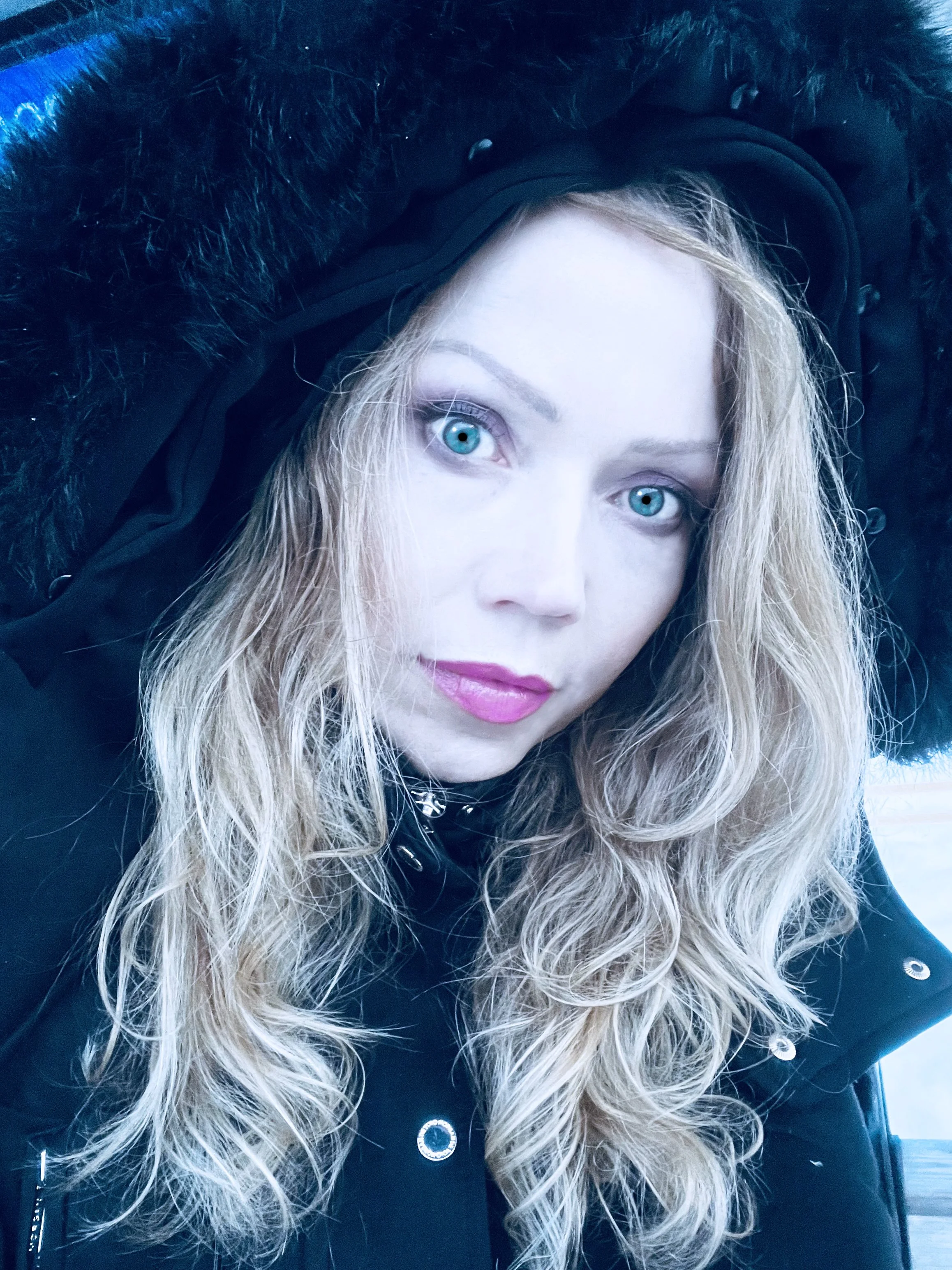 Close-up of a woman with long curly blonde hair, blue eyes, wearing a black jacket with fur trim, looking directly at the camera.