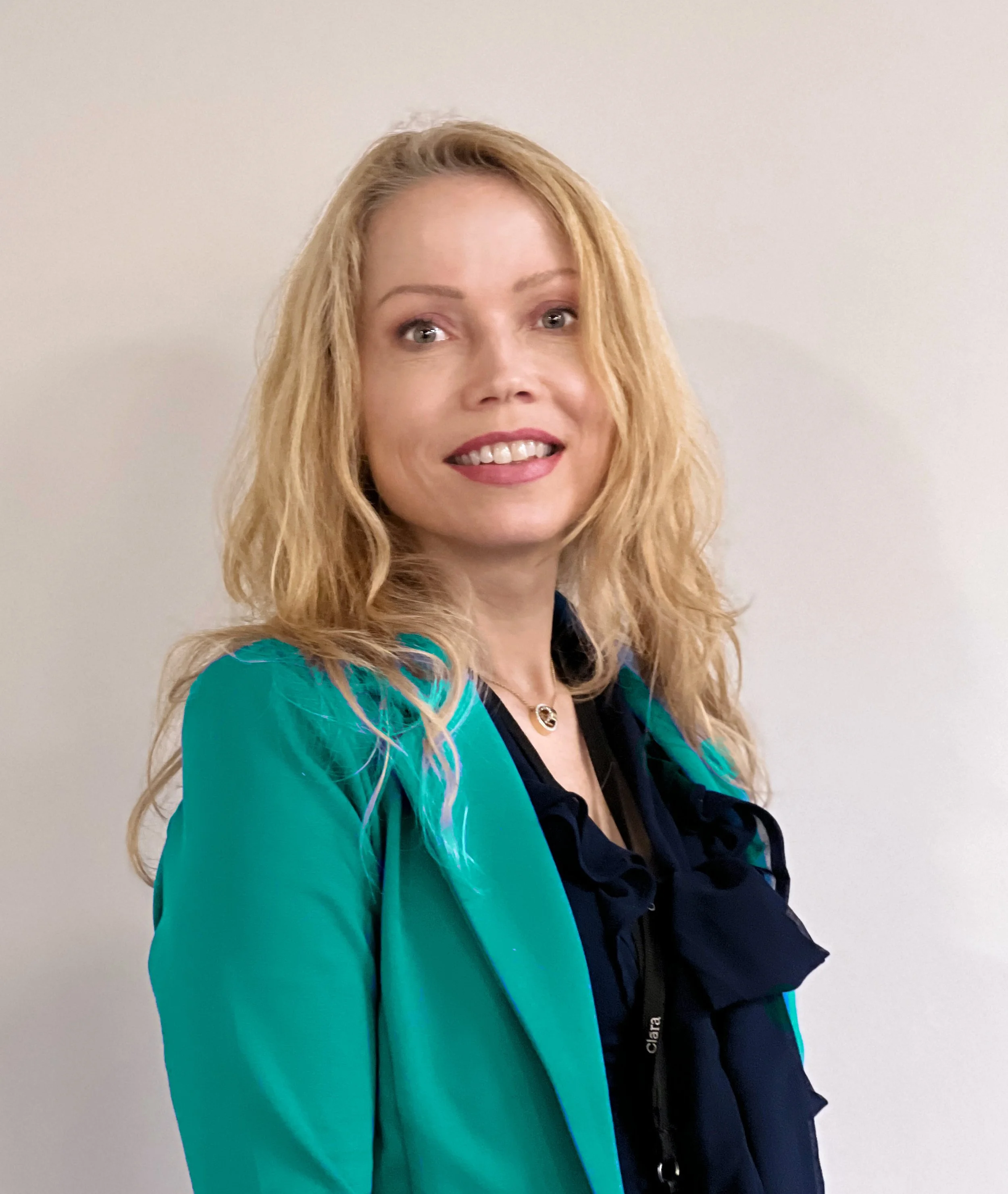 Portrait of a smiling blonde woman with wavy hair, wearing a teal blazer over a black top, standing in front of a plain light-colored wall.