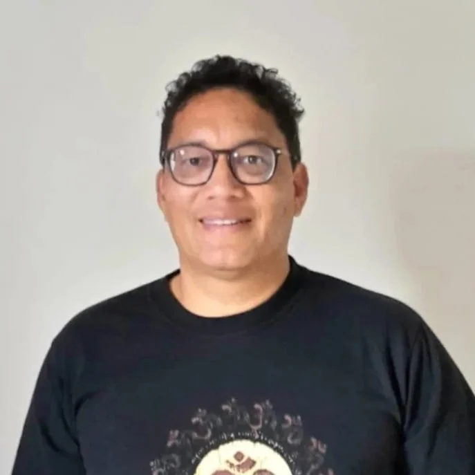 A man wearing glasses and a black t-shirt standing in front of a plain light-colored wall.