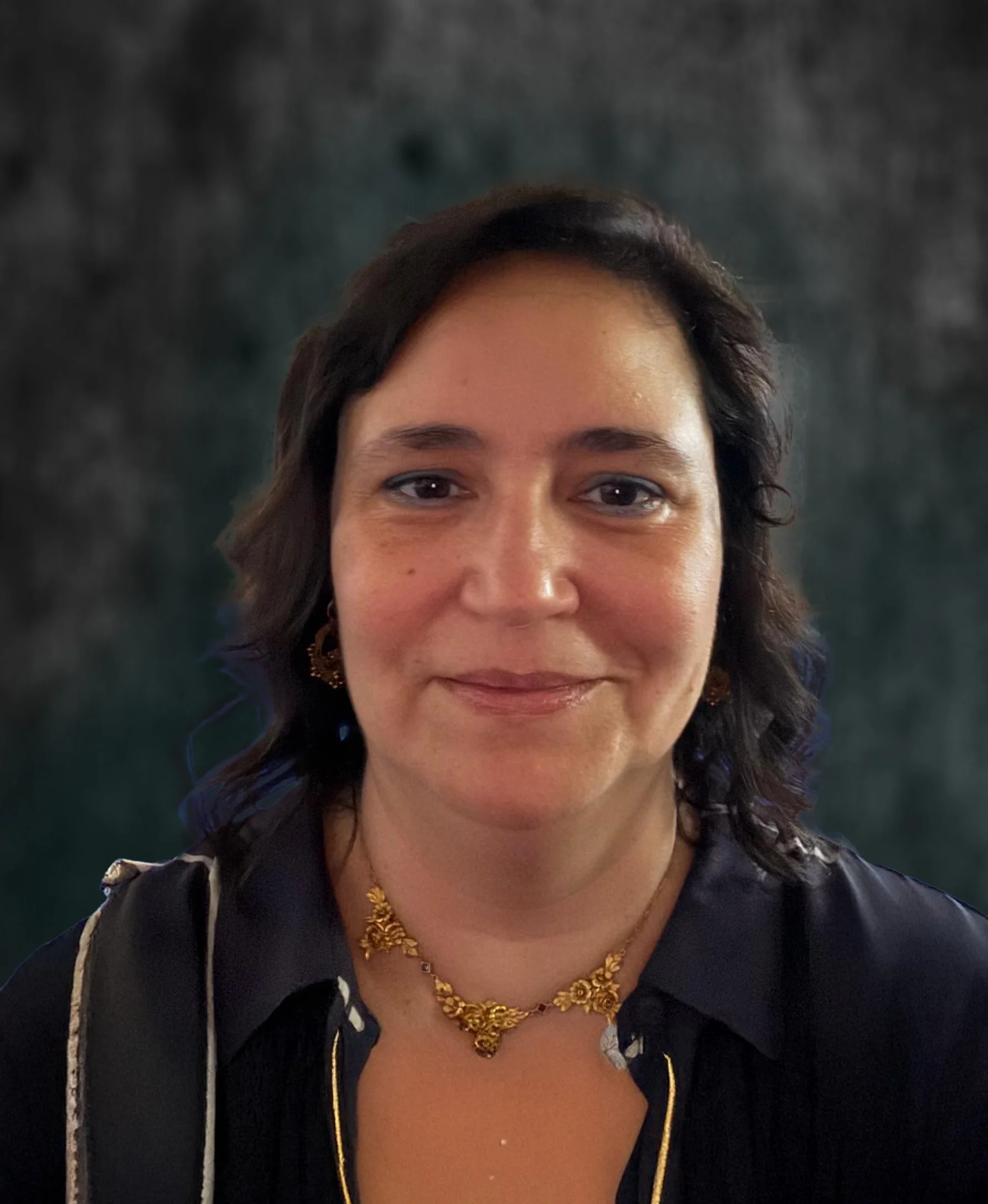Close-up of a woman with dark hair wearing a gold necklace and earrings, against a dark background.