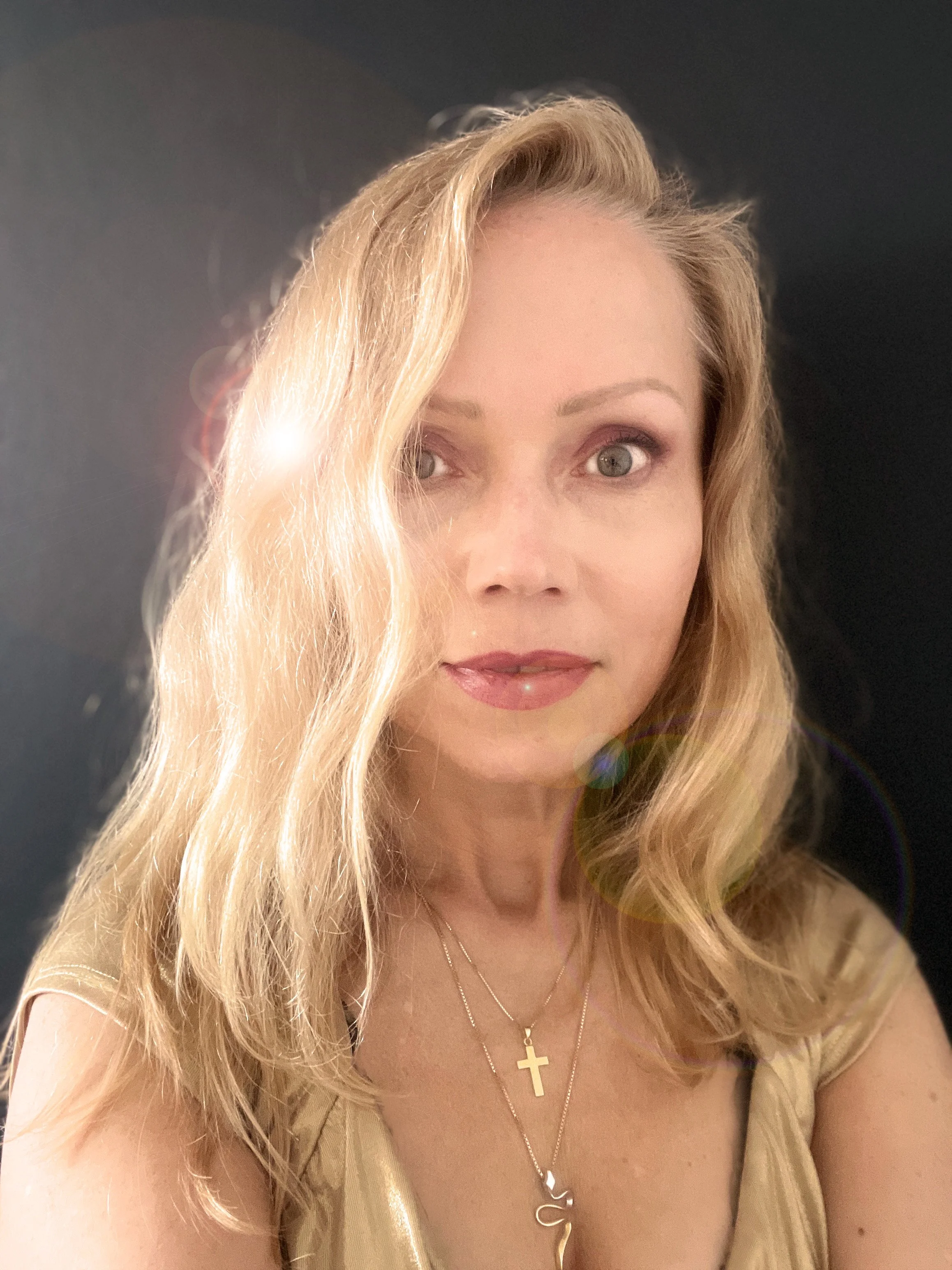 Close-up of a woman with blonde hair wearing layered necklaces, including a cross and letter 'U' pendant, against a dark background.